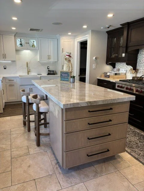 Modern kitchen with white cabinets, a beige island, black bar handles, two bar stools, a stainless steel stove, and dark upper cabinets.