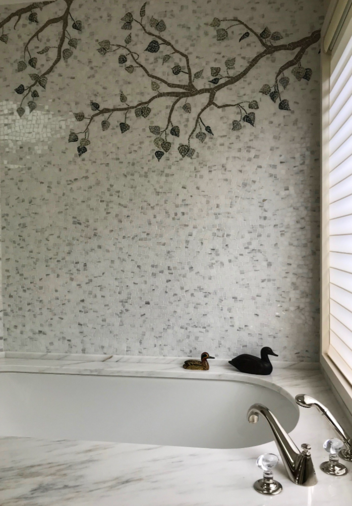 Bathroom with a marble bathtub, a mosaic tile wall featuring a tree branch with leaves, and two rubber duckies on the tub's edge, near a window with blinds.