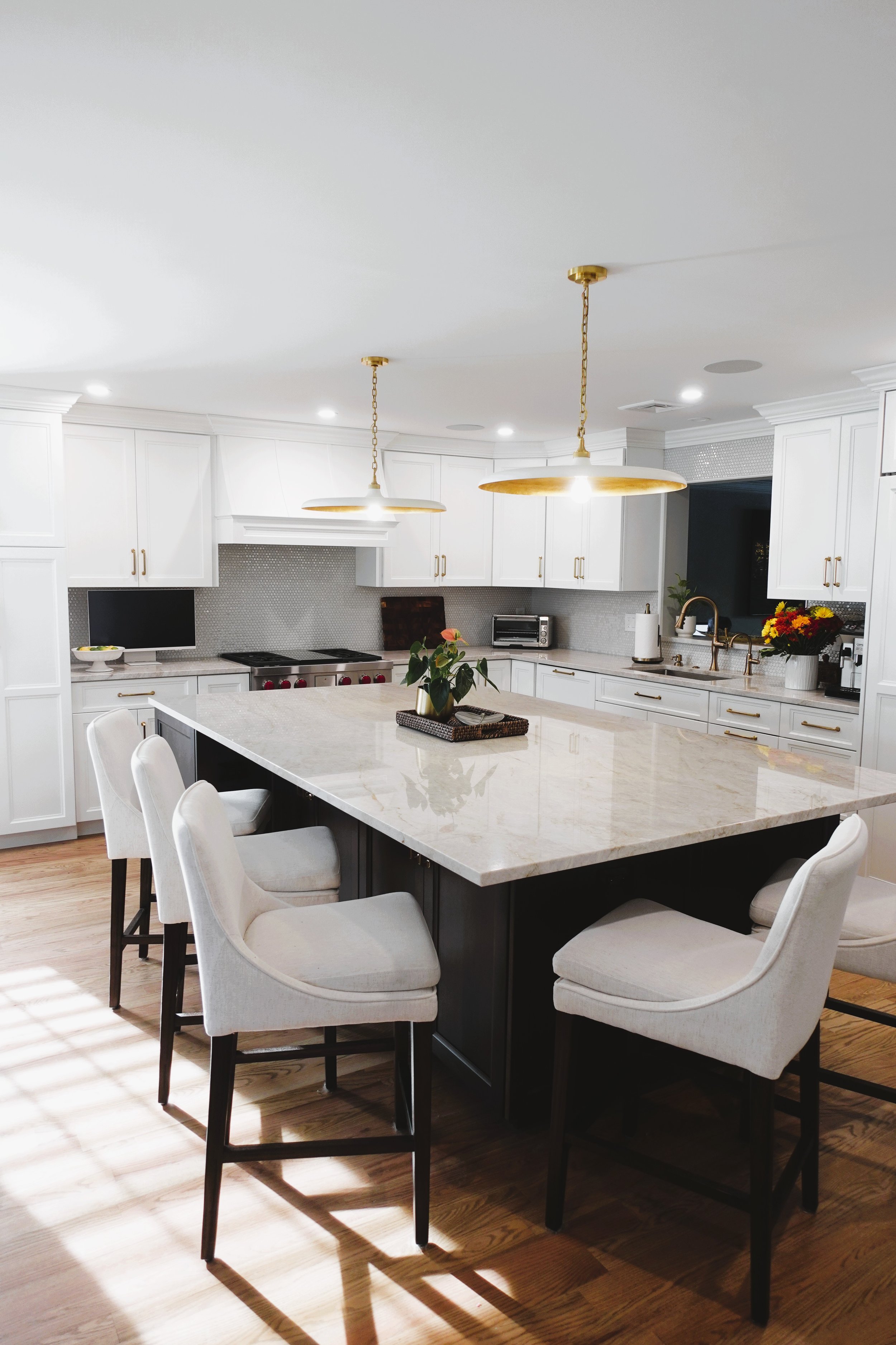 Modern kitchen with white cabinets, gold handles, and a large marble island surrounded by white chairs. Gold pendant lights hang over the island, and there are plants and flowers on the countertop, with a window and a small television in the background.