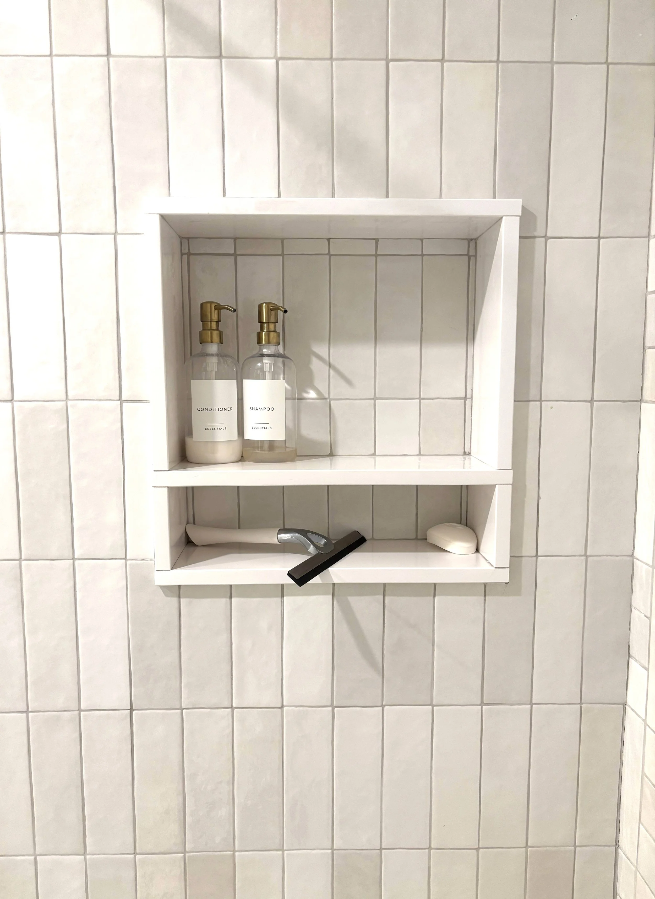 A bathroom shower shelf with two bottles labeled conditioner and shampoo, a razor, a bar of soap, and a black hairbrush, all mounted on white tiled wall.