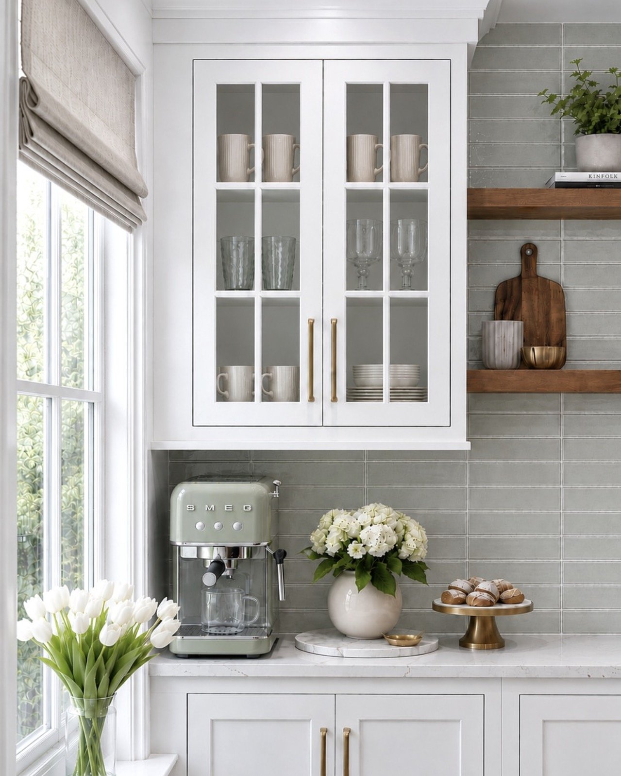 A kitchen corner with white cabinets, glass door, and a marble countertop. Inside the open cabinet are beige mugs, glassware, and stacked plates. A Smeg espresso machine, a vase with white flowers, and a tray of chocolates are on the counter. A window with a beige Roman shade is on the left, and wooden shelves with decorative items are on the right.