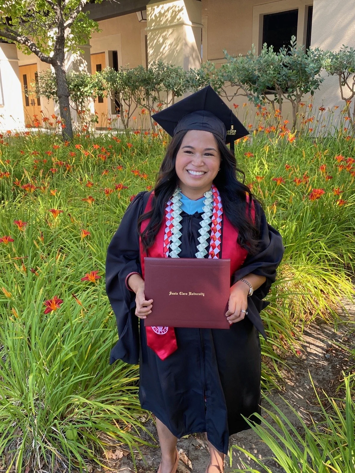 Woman in graduation cap and gown holding a diploma cover in a garden with orange flowers and green bushes.