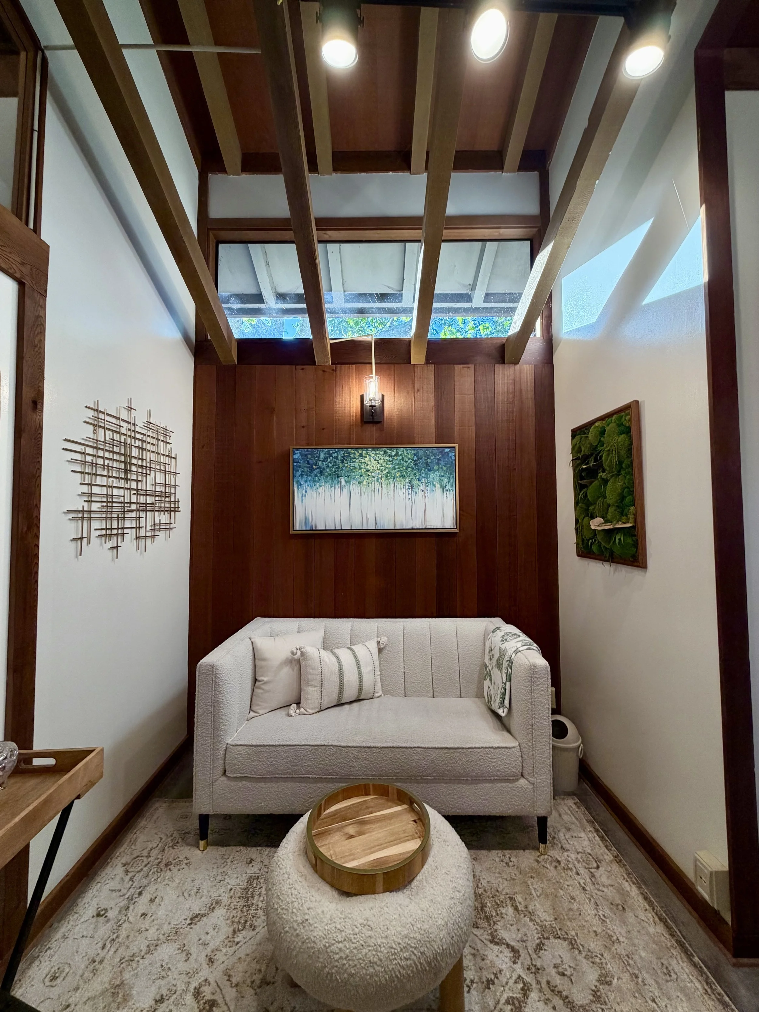 A cozy living room with a white upholstered armchair, a round textured ottoman with a wooden tray on top, artworks on the wall, and a wooden ceiling with skylights.