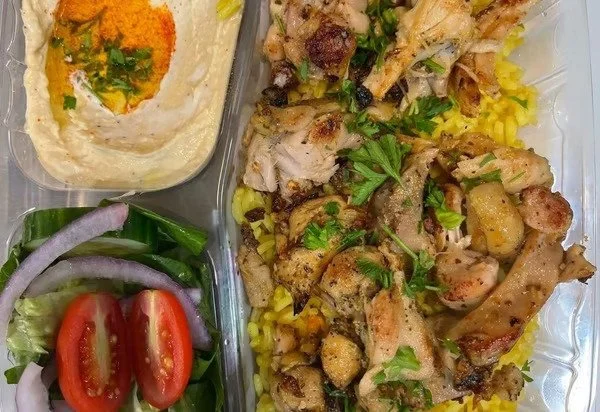 Plate of grilled chicken served over yellow rice, a side of fresh salad with cherry tomatoes, cucumber, and onion slices, and a serving of hummus garnished with paprika and parsley.