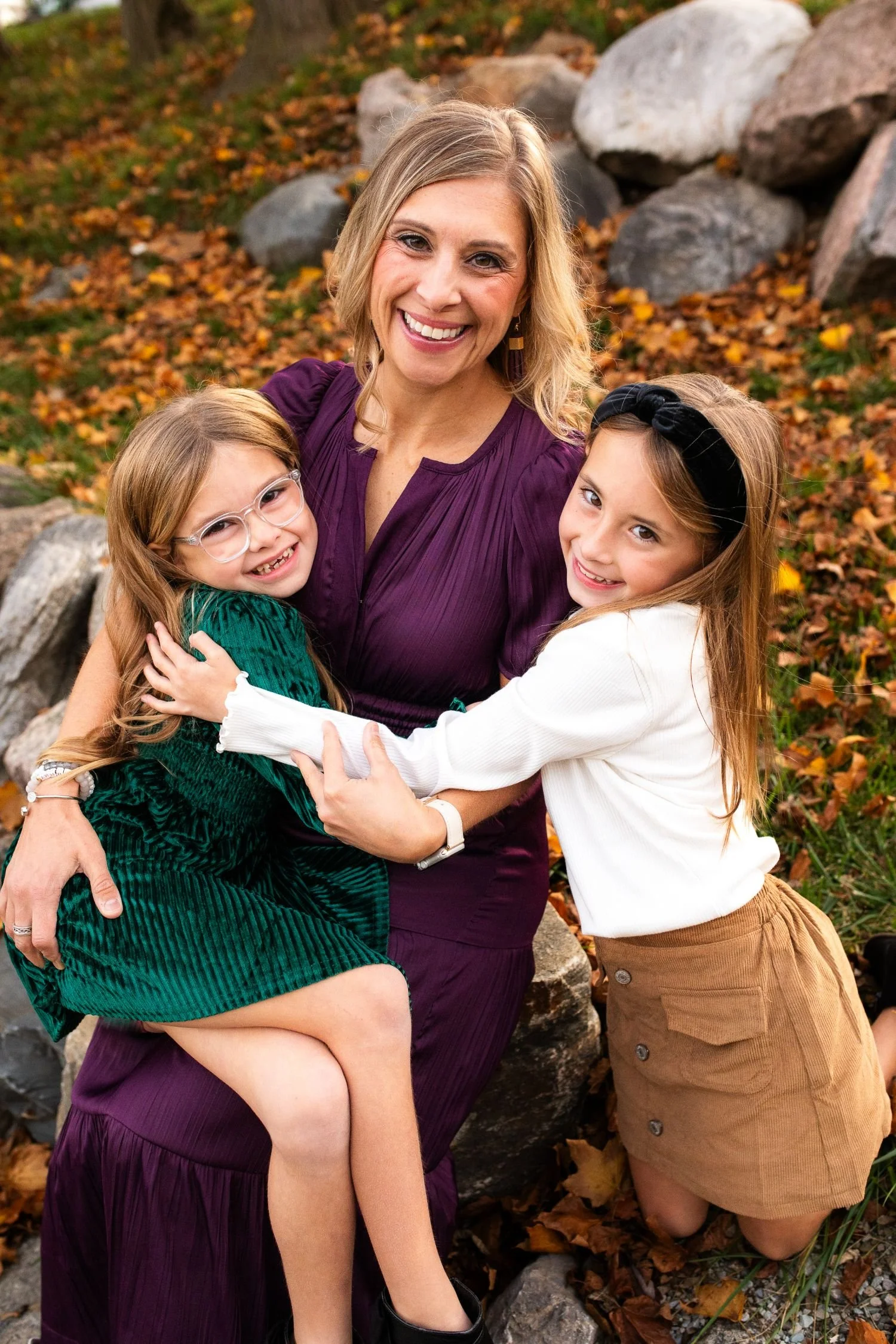 Mother in a purple dress smiling as she hugs her two young daughters outdoors, surrounded by rocks and colorful autumn leaves.