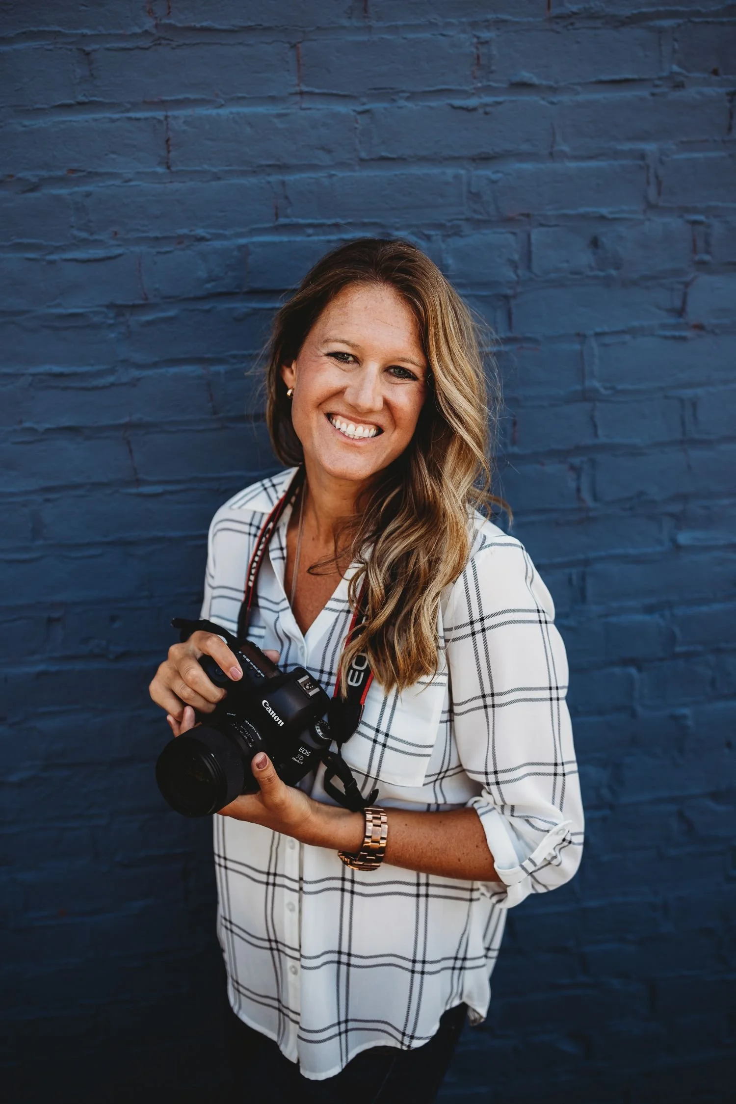 Smiling female photographer with long blonde hair wearing a white plaid shirt and holding a professional DSLR camera in front of a bright blue brick wall.