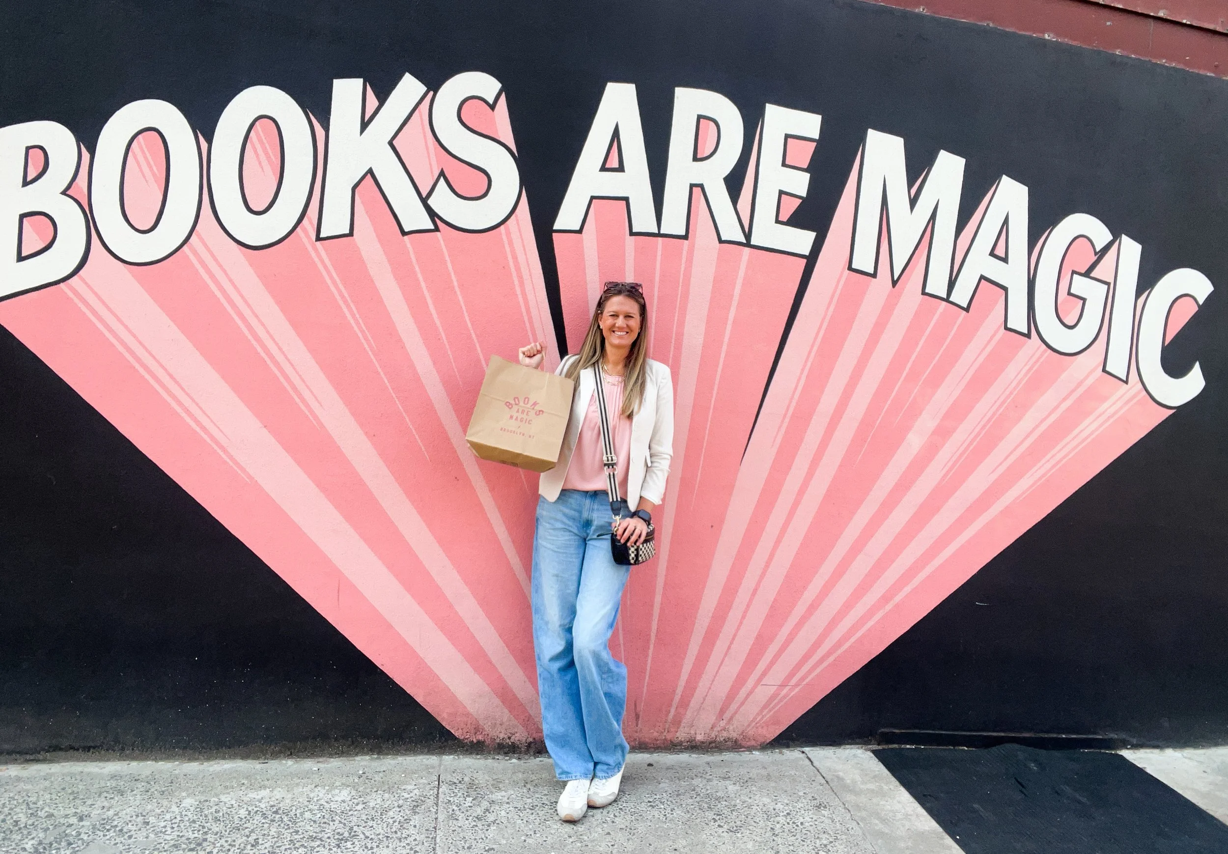 Smiling woman wearing a white jacket, light blue jeans, and sneakers, holding a shopping bag and camera while standing in front of a colorful pink and black mural that reads "Books Are Magic."