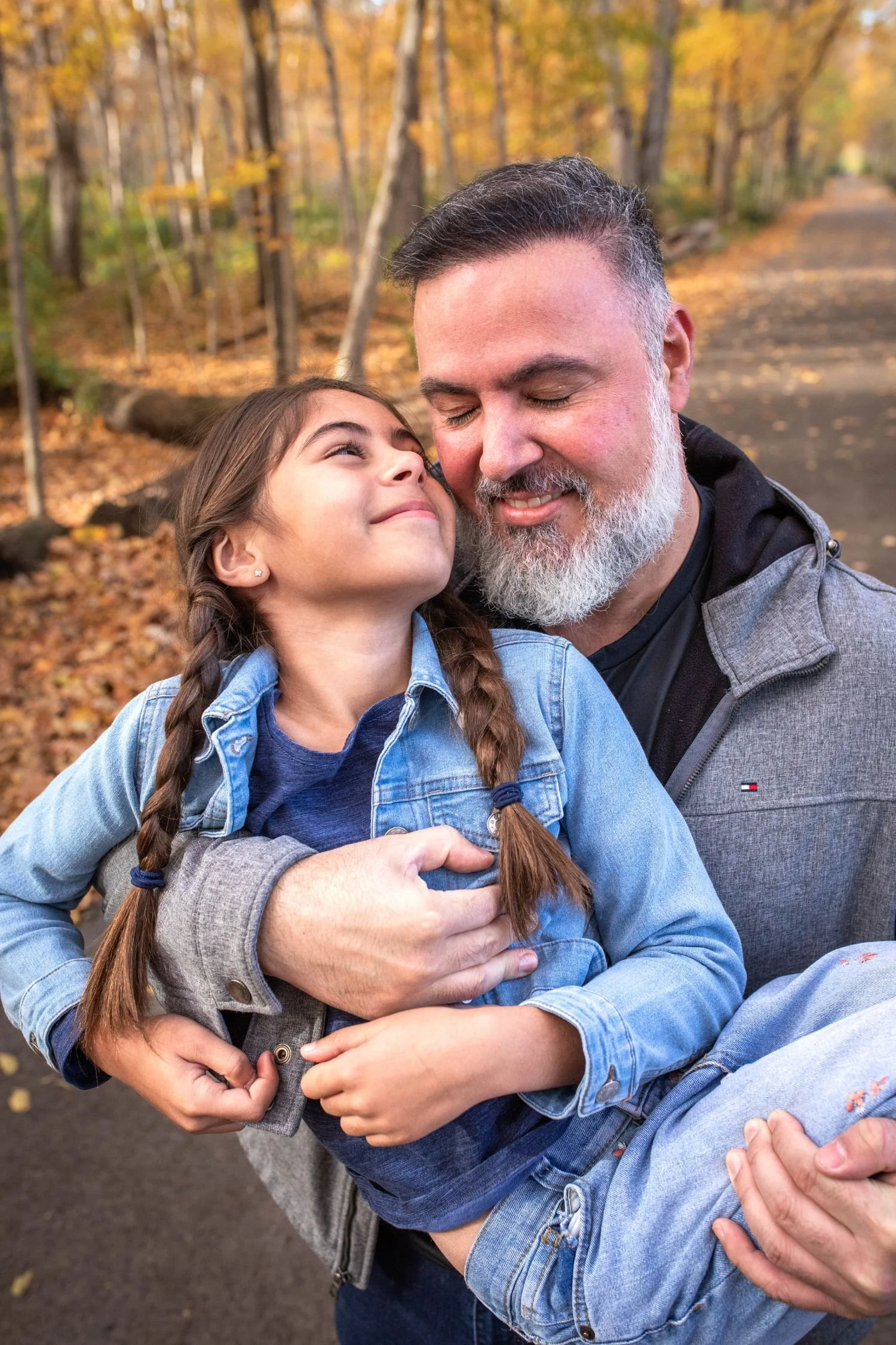Father smiling as he hugs his young daughter with braided hair and a denim jacket on a wooded path surrounded by colorful fall foliage.