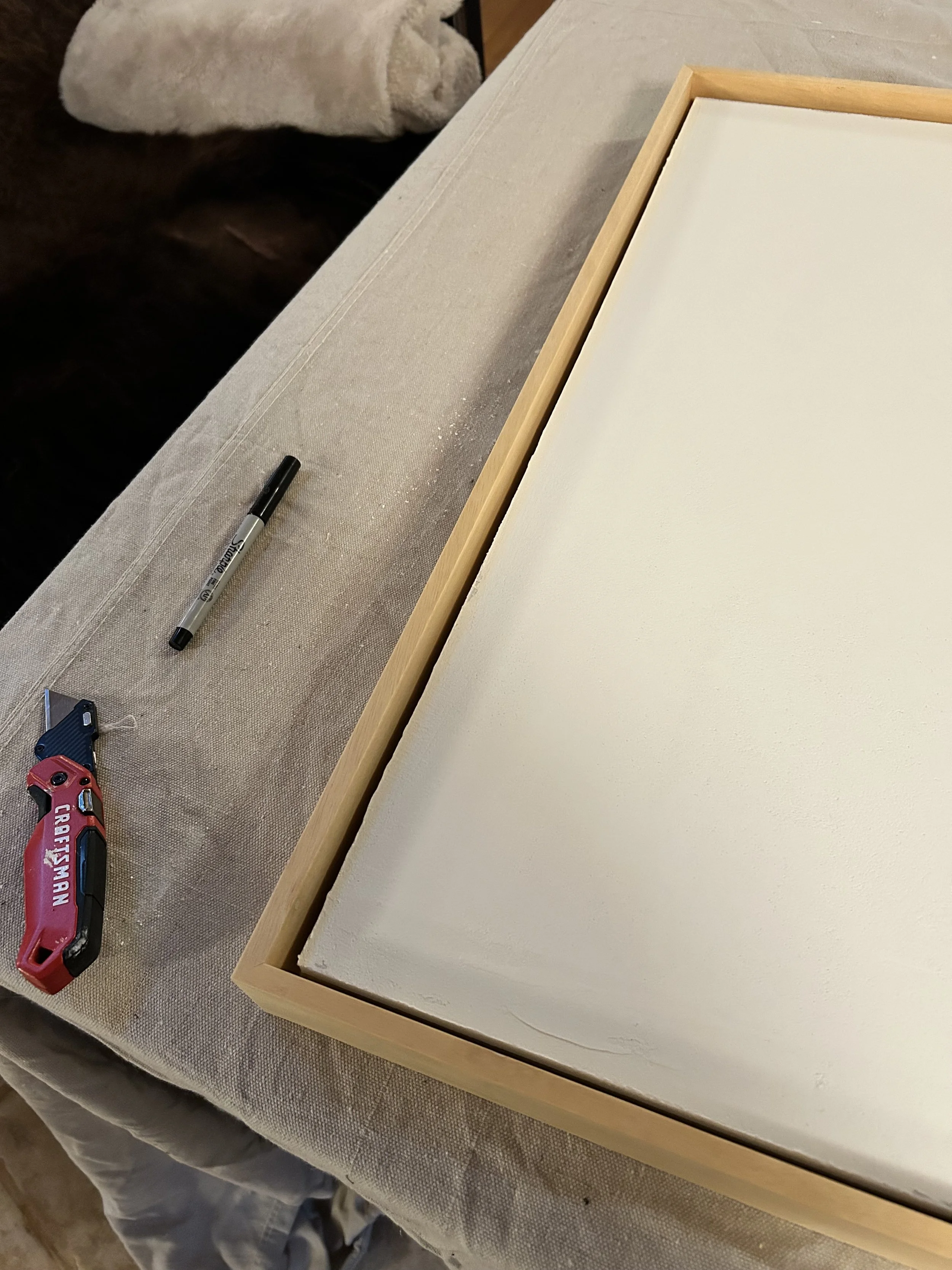 A wooden frame on a table with a white surface inside it. There is a black Sharpie marker, a red and black Craftsman cordless drill, and a black laser level on the table. The table is covered with a beige tablecloth.