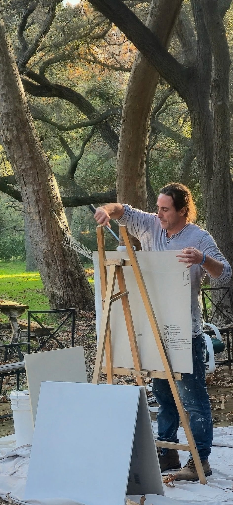 An artist painting outdoors on an easel surrounded by trees, with empty canvases and art supplies nearby.
