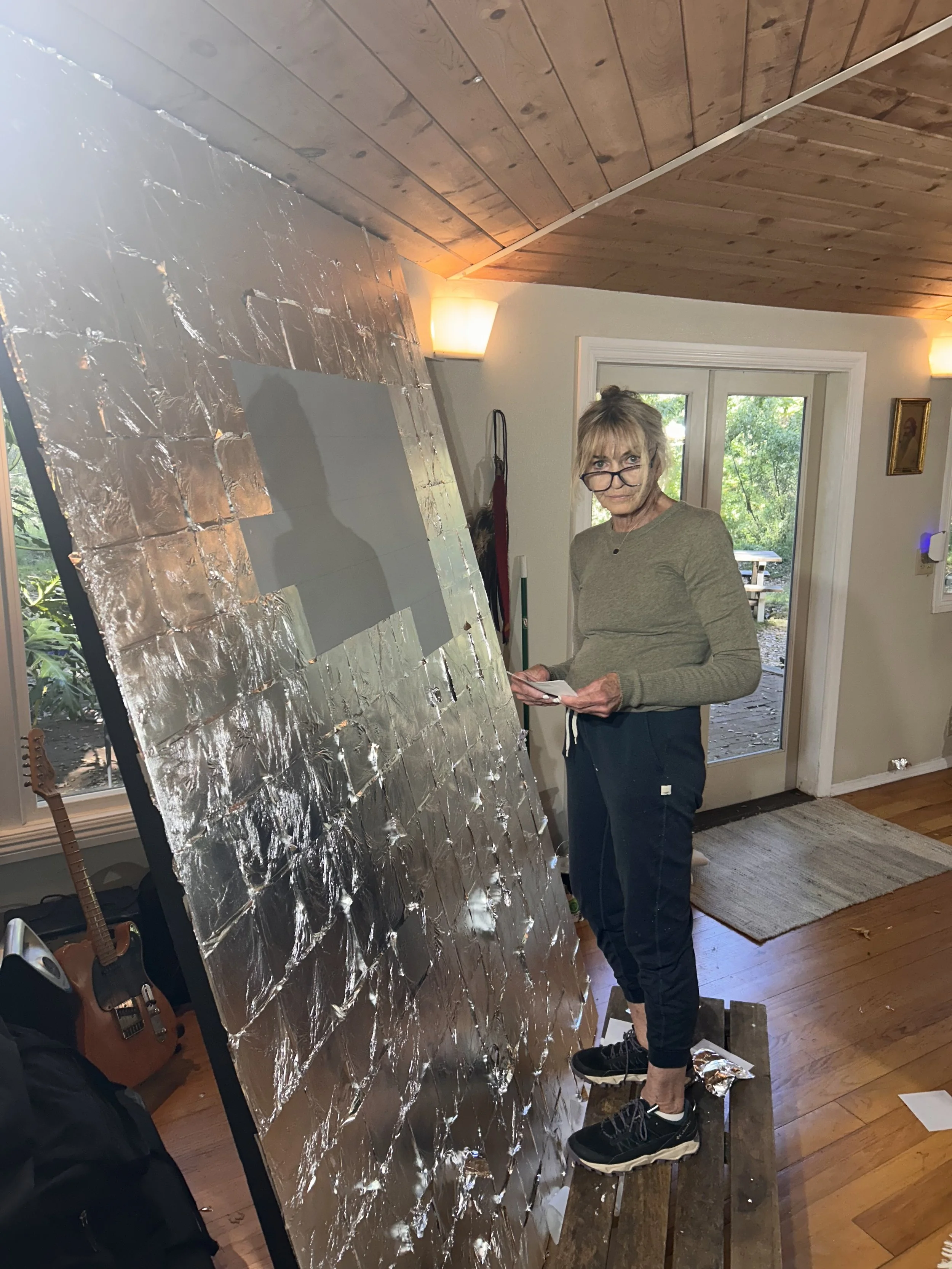 An elderly woman with glasses, wearing a gray sweater and black pants, standing inside a home near a large rectangular sheet of reflective material, possibly foil, which is partially covering a surface.