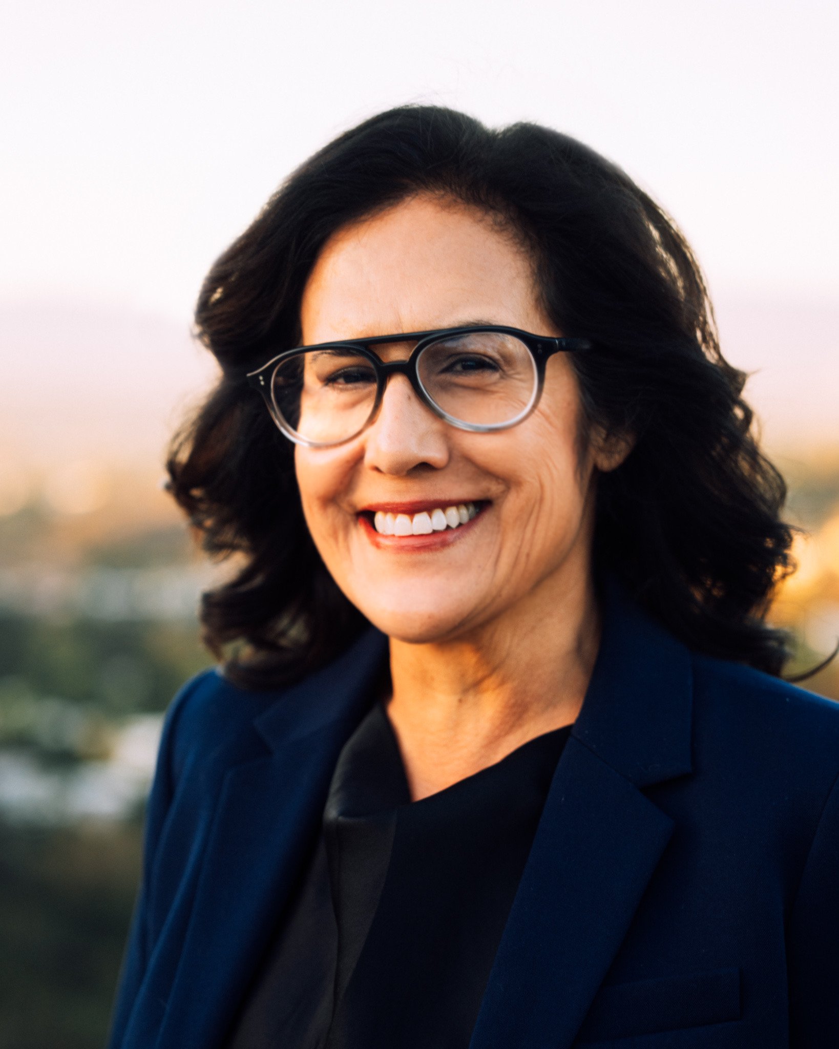 Headshot of Liz Morentin, a smiling woman with dark curly hair, glasses, wearing a dark blue blazer, standing outdoors during sunset.