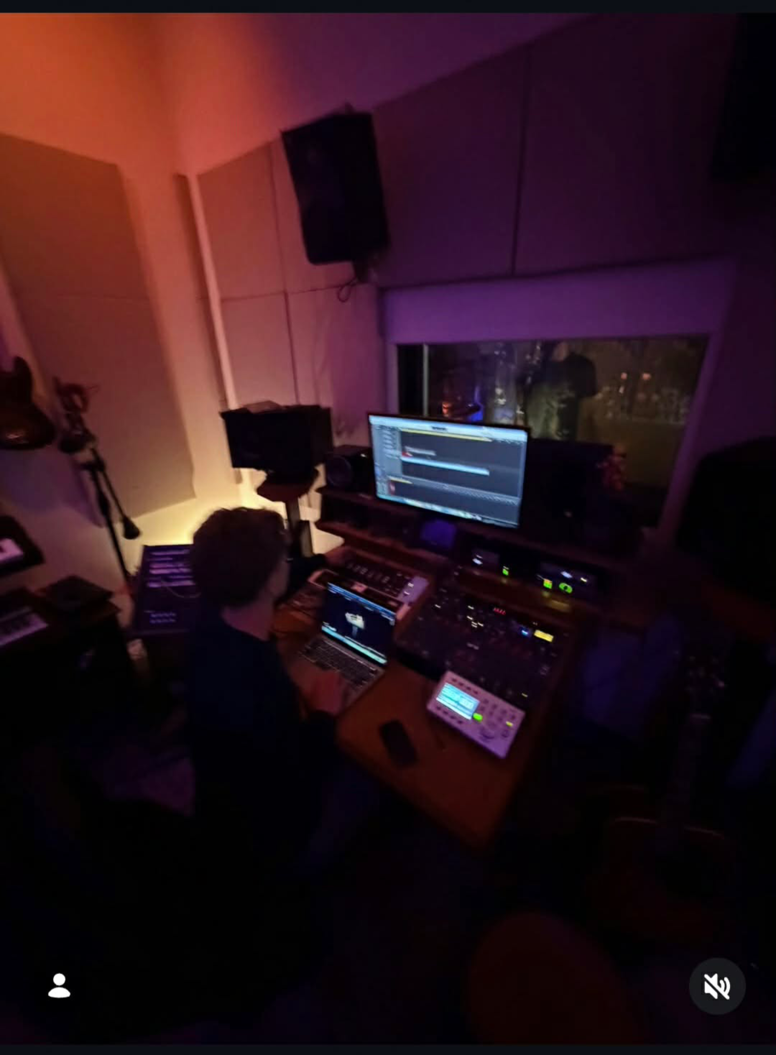 A home recording studio with a computer monitor displaying music production software, studio monitors, a keyboard, and a person working at the desk in a dimly lit room with purple and orange lighting and a window showing a nighttime city view.