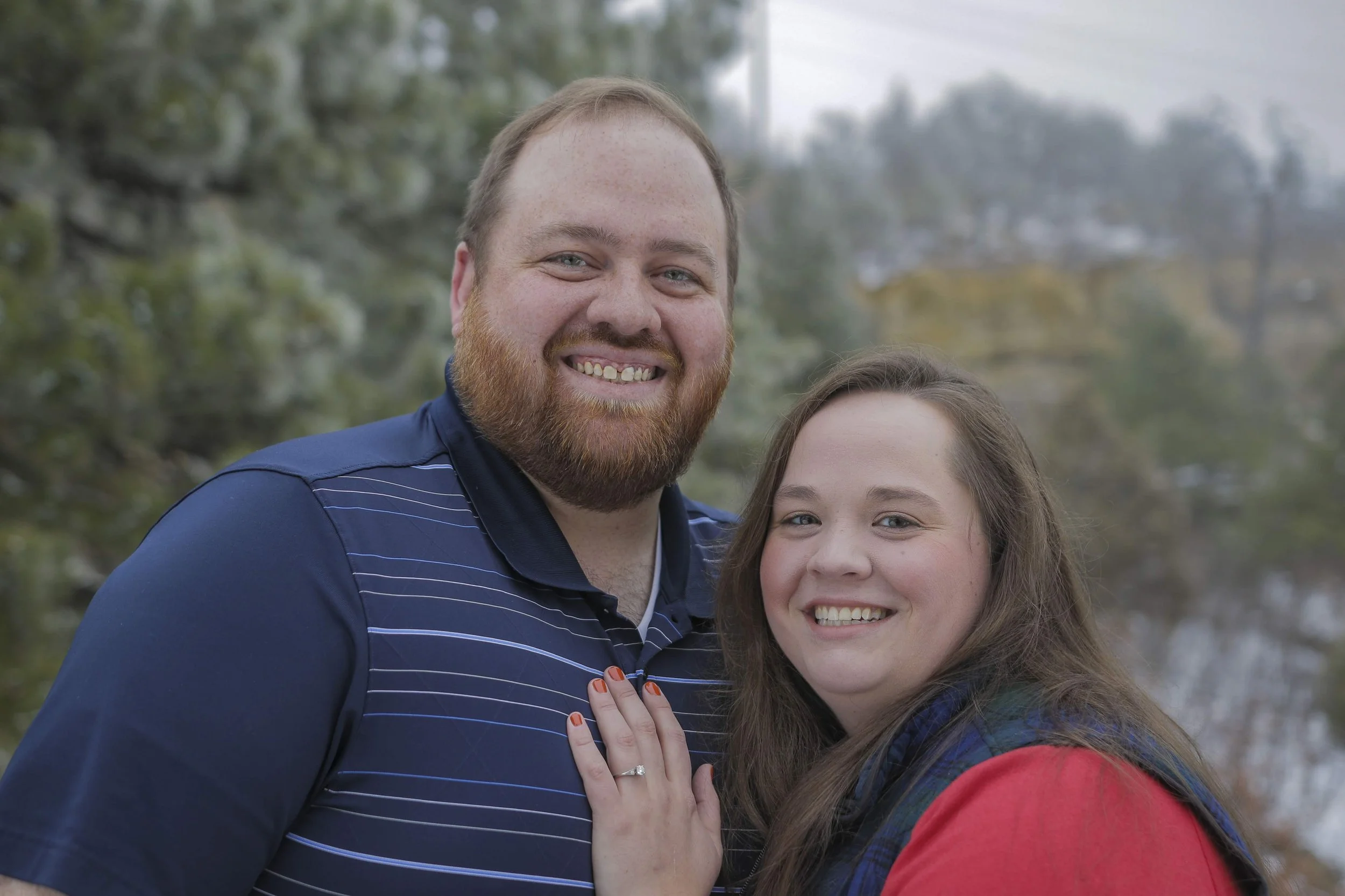 Photo of owners, Mike and Jenny Darnell. Happy man with red hair and beard wearing a dark blue striped polo shirt standing outdoors next to a young woman with long brown hair wearing a red jacket and a blue scarf, both smiling.