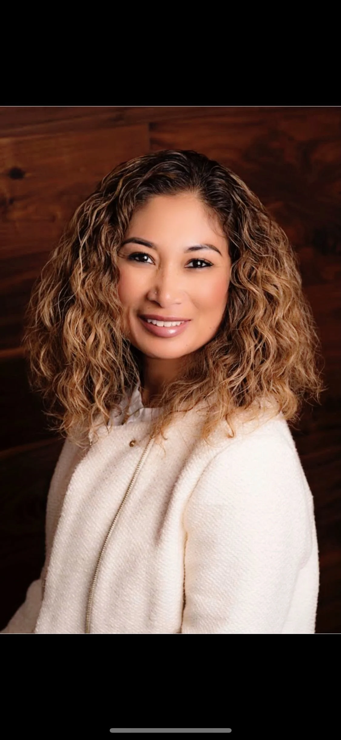 Portrait of a woman with curly light brown hair, wearing a white jacket, smiling in front of a wooden background.