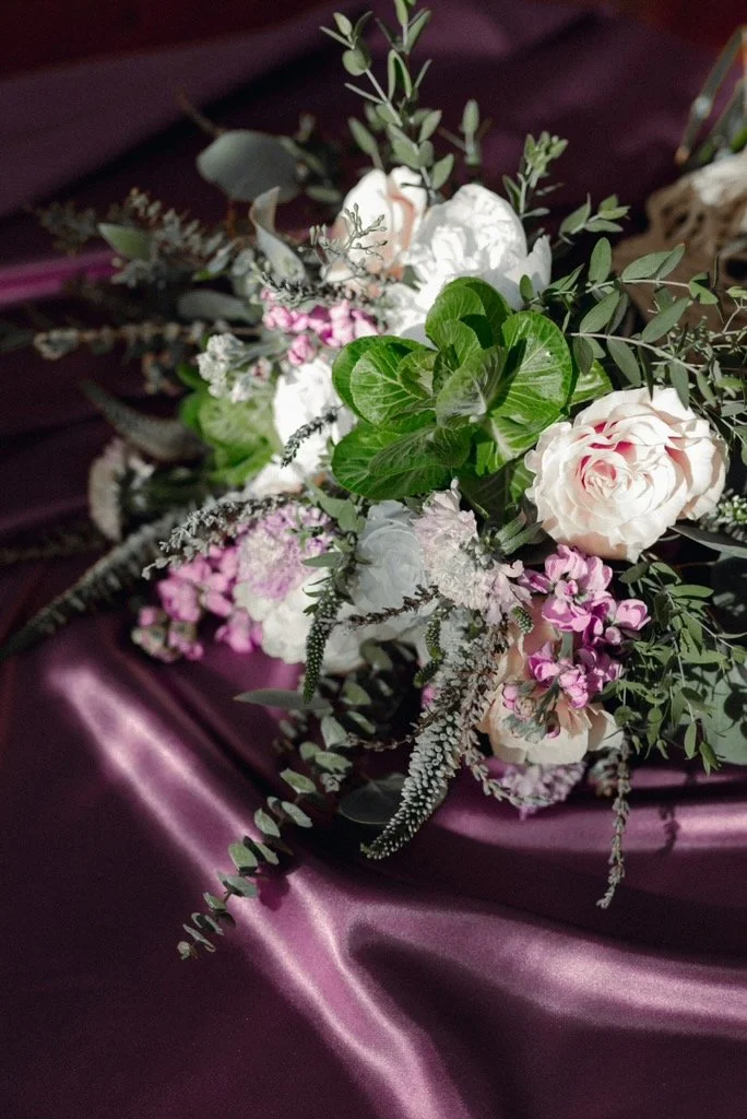 A floral arrangement featuring white roses, pink flowers, and greenery placed on a dark purple satin fabric.
