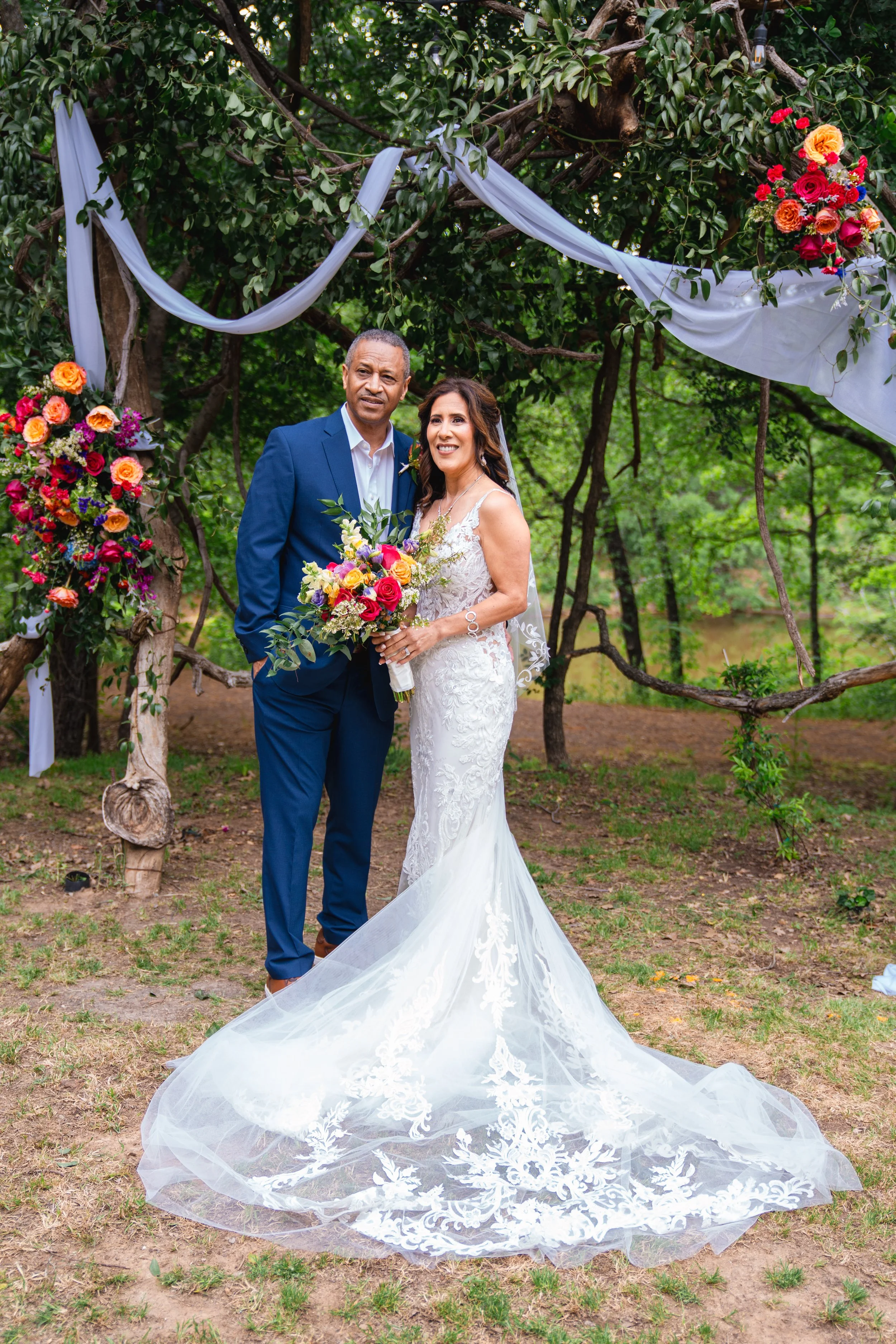 A couple dressed in wedding attire standing outdoors under a decorated tree, with the bride holding a bouquet of colorful flowers.