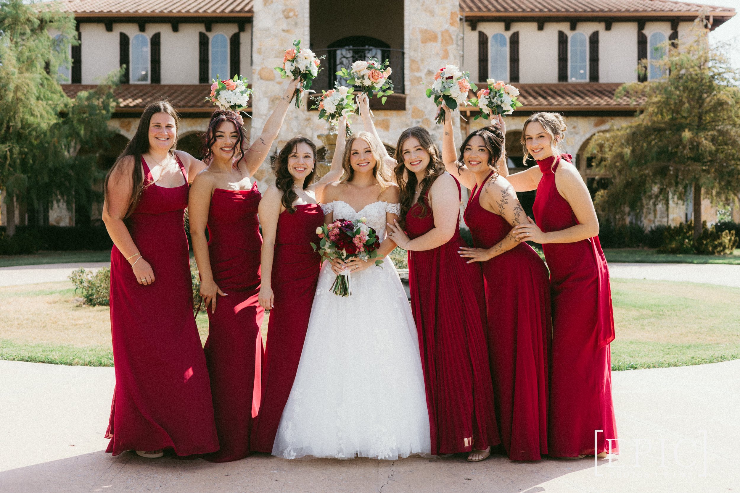 Group of seven women, including a bride in a white wedding dress holding a bouquet, celebrating with six bridesmaids dressed in matching red gowns, outdoors in front of a stone building with trees nearby.