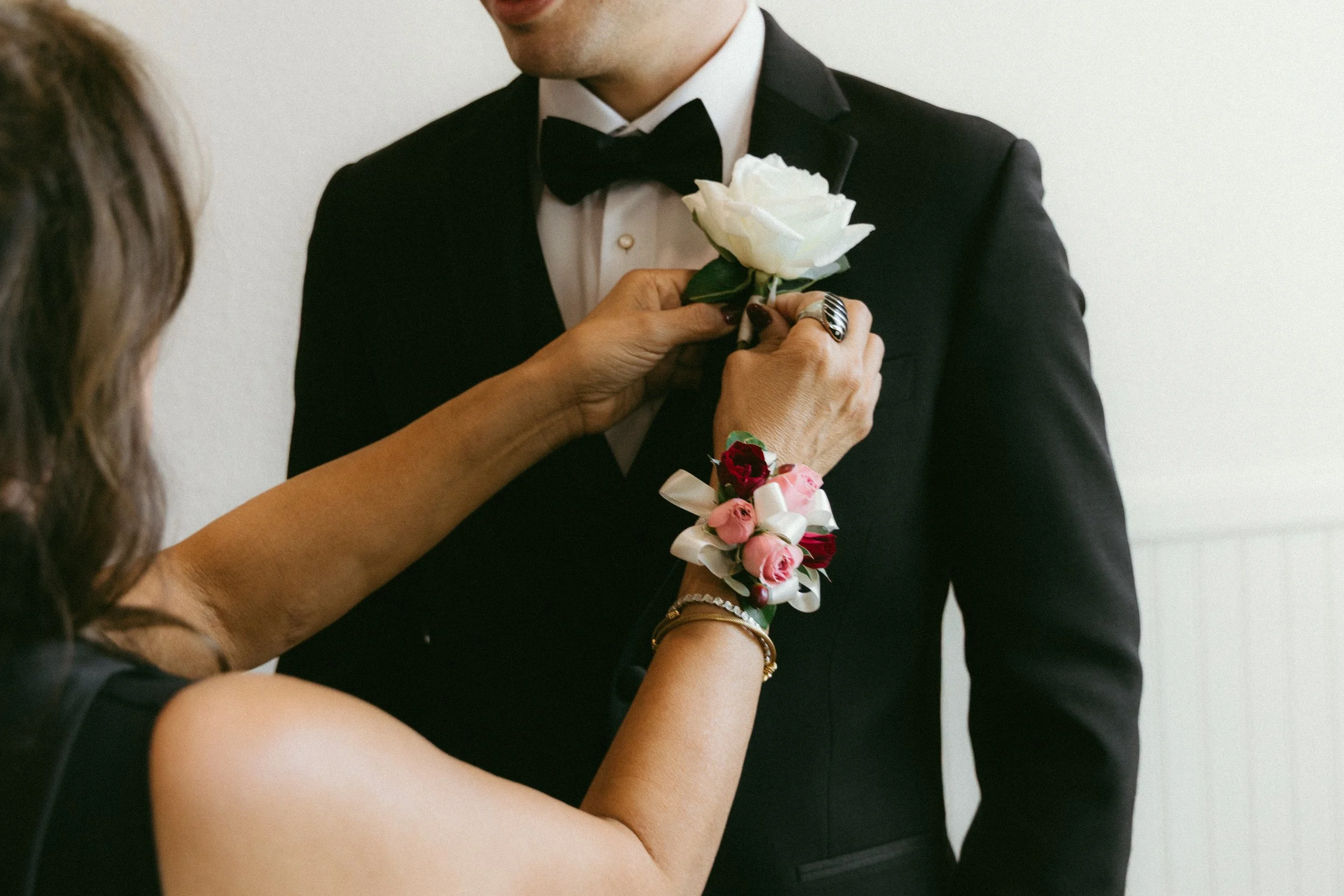 A person in a tuxedo with a black bow tie is receiving a white rose from another person, who is attaching a floral corsage with pink and red roses to their suit.