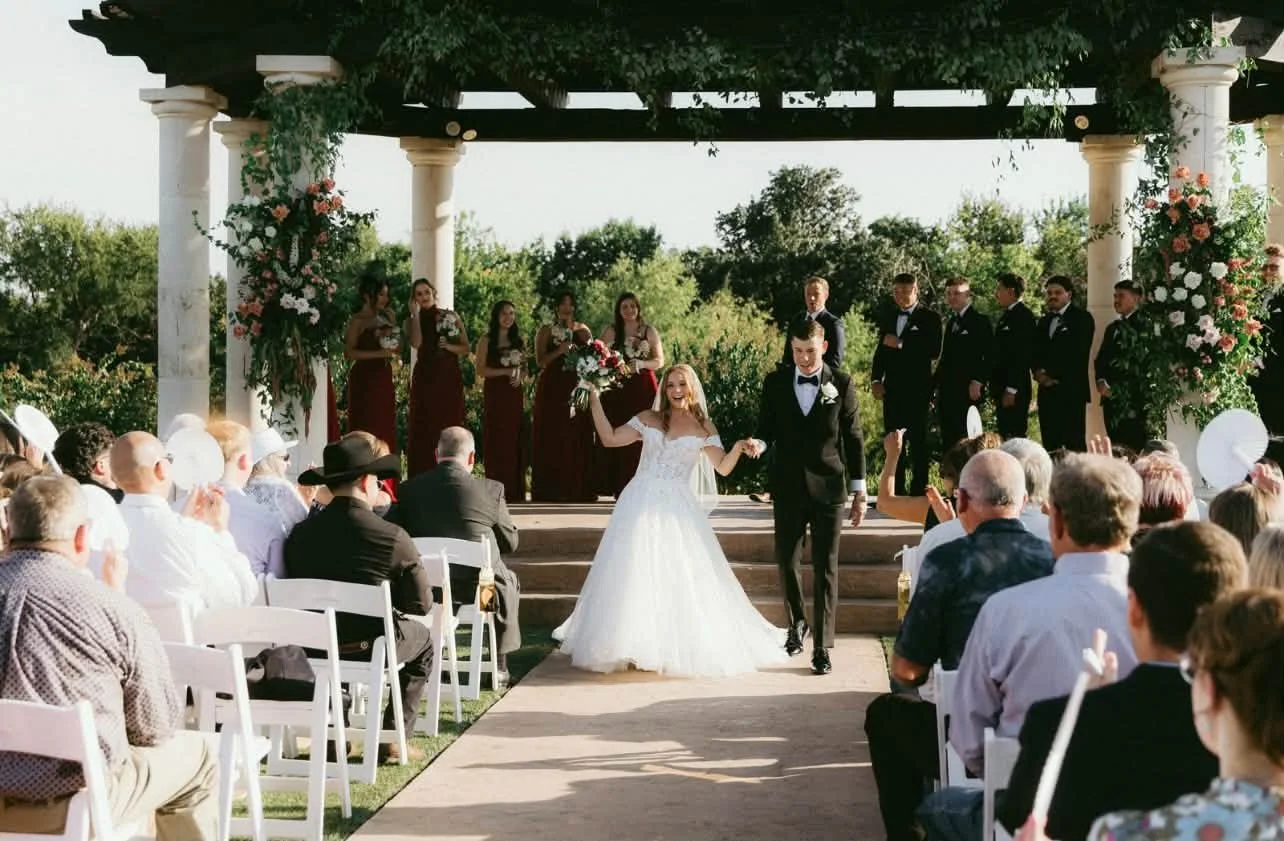 Outdoor wedding ceremony with a bride and groom walking down the aisle, surrounded by seated guests and a wedding party on the stage, decorated with flowers and greenery.