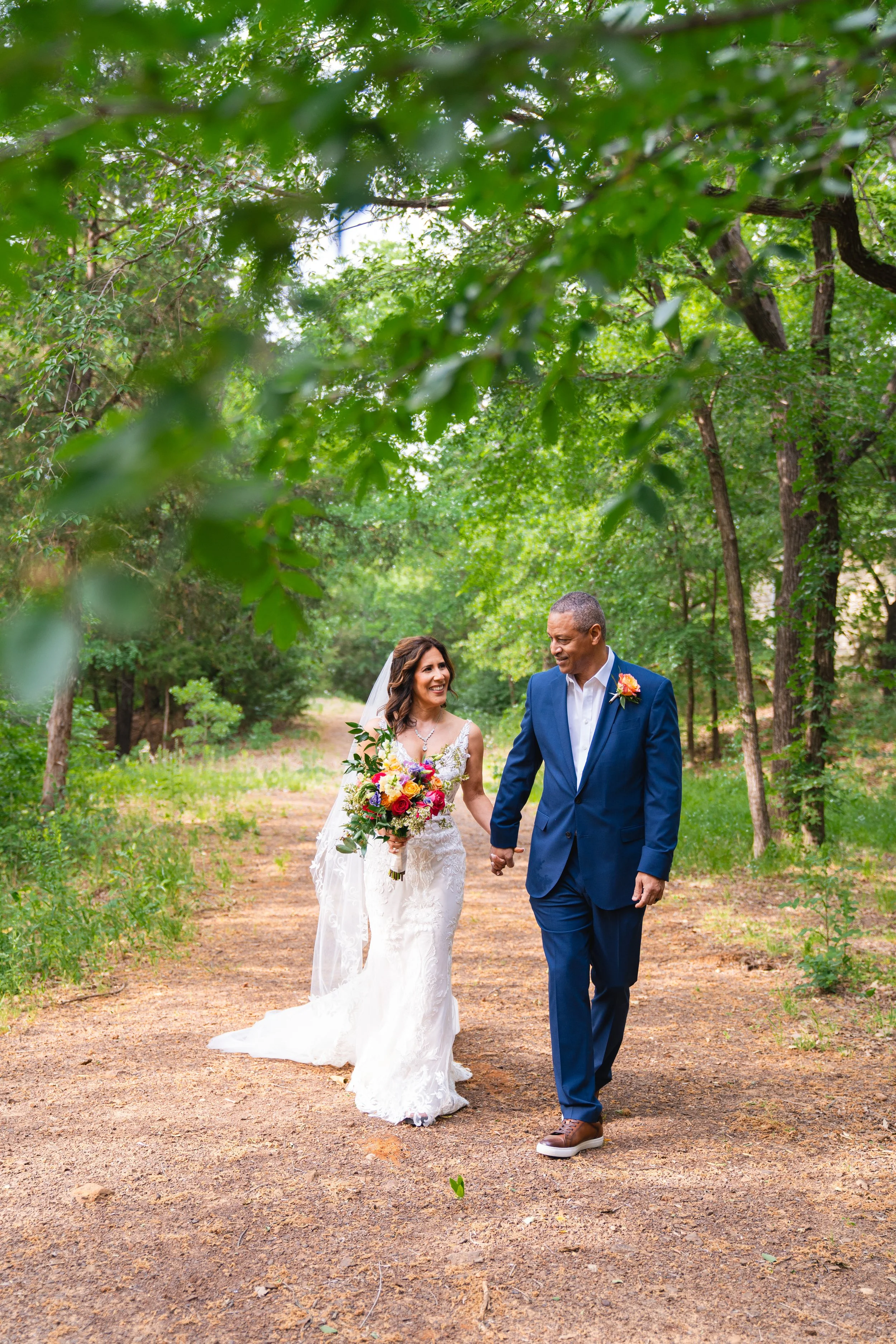 A newly married couple walking hand in hand on a dirt path through a green wooded area. The bride is wearing a lace white wedding dress and holding a colorful bouquet. The groom is dressed in a blue suit and brown shoes, and both are smiling happily.