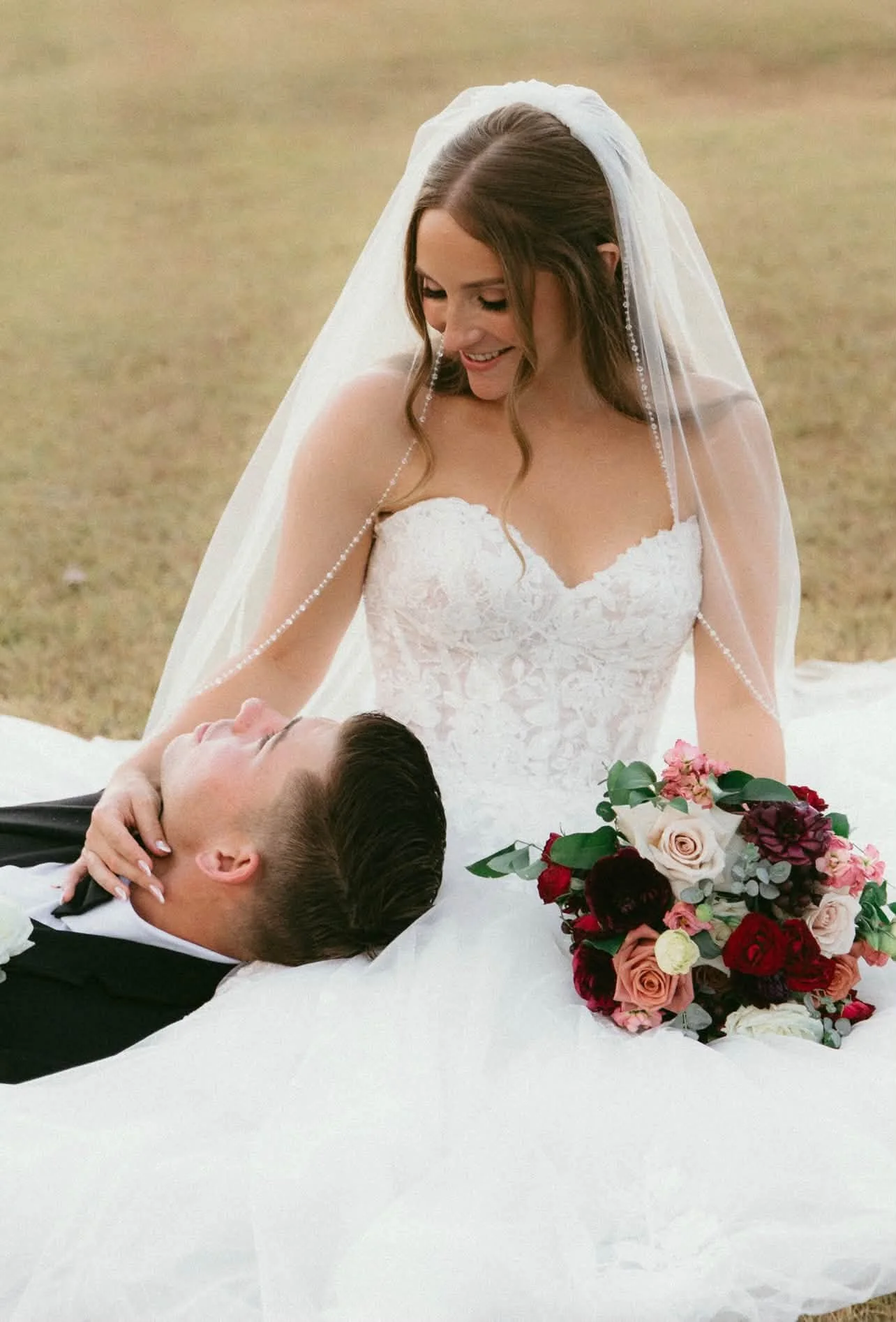A bride in a white lace wedding dress and veil, smiling down at a groom lying on a white surface, with a bouquet of roses and greenery nearby, outdoors.
