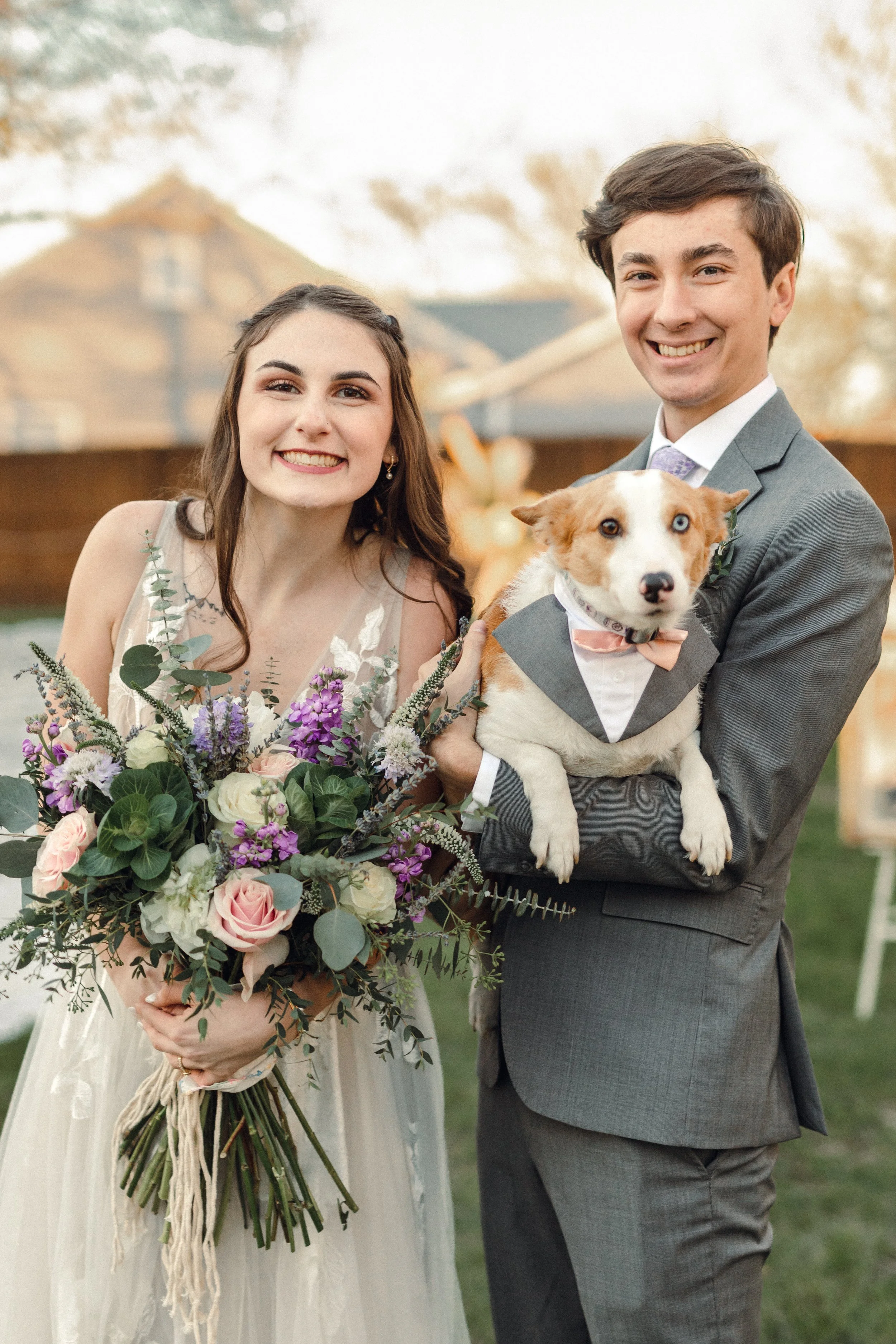 A smiling bride in a white dress holding a large bouquet of pink, purple, and white flowers. Standing next to a smiling groom in a gray suit holding a dog with heterochromatic eyes (one blue, one brown), dressed in a tuxedo with a bow tie. They appear to be outdoors at a wedding.