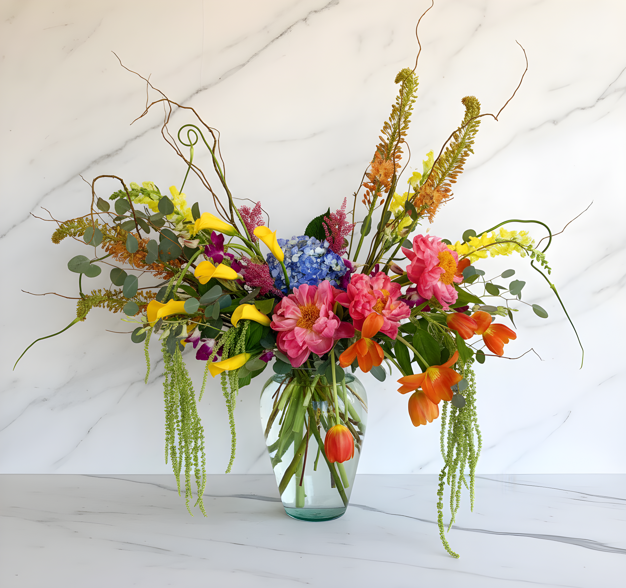 A colorful floral arrangement in a glass vase on a white marble surface, featuring pink peonies, orange tulips, yellow flowers, purple orchids, green leaves, and twisting branches with a minimalist background.