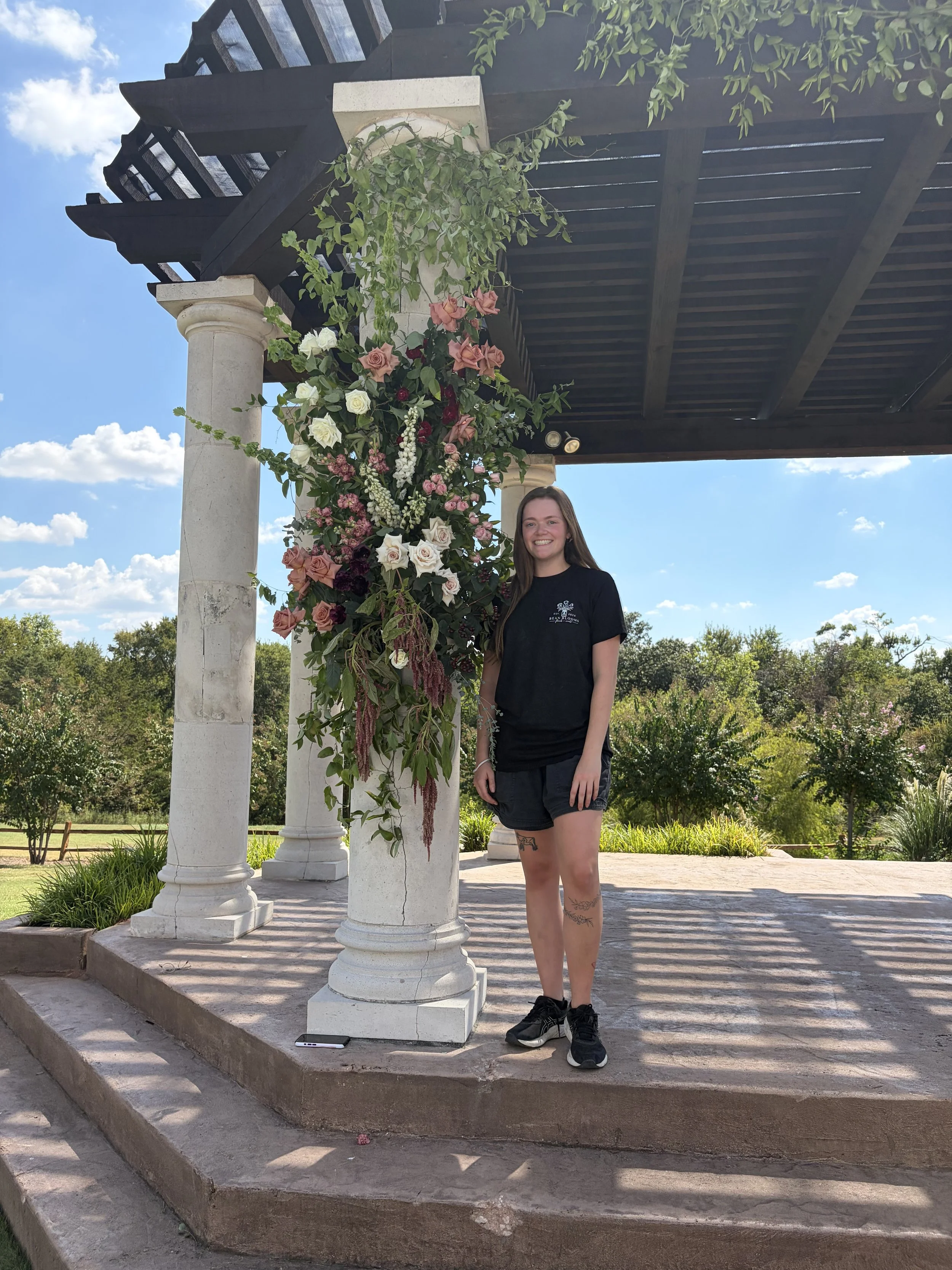 A young woman with long hair, wearing a black t-shirt, shorts, and sneakers, standing next to a stone pillar decorated with a large floral arrangement of roses and greenery under a wooden pergola in an outdoor park setting with trees and blue sky.