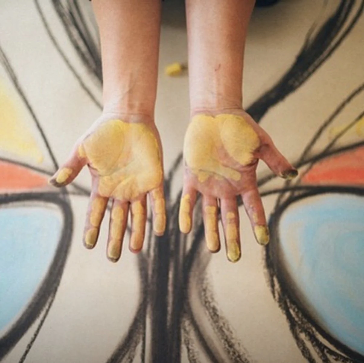 A person’s hands covered in yellow paint, with a black abstract artwork in the background.