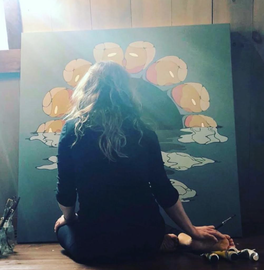 An artist with long hair sitting cross-legged on the floor in front of a large painting of a lotus flower reflected in water, with art supplies nearby.