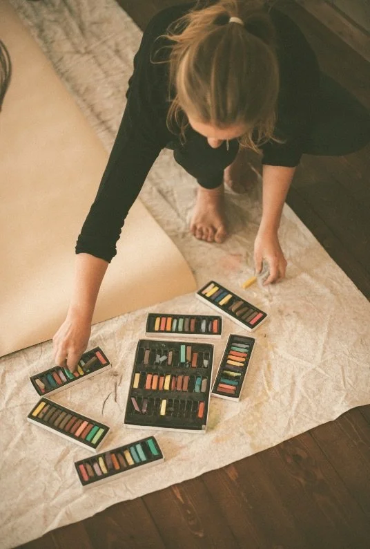 Person organizing colorful pastels and chalks on a large sheet of paper on the floor