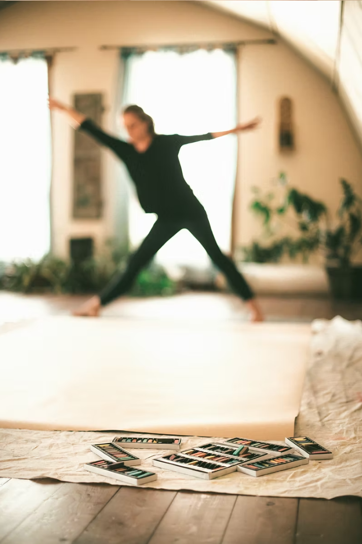 A woman practicing yoga in a bright room with large windows, with art supplies, such as pastels or colored pencils, spread on the floor in the foreground.