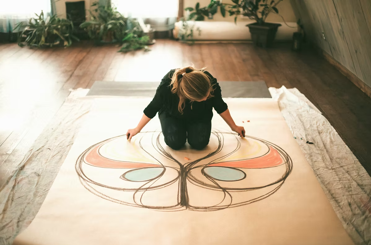 A woman kneeling on the floor and drawing a colorful butterfly design on a large piece of paper.