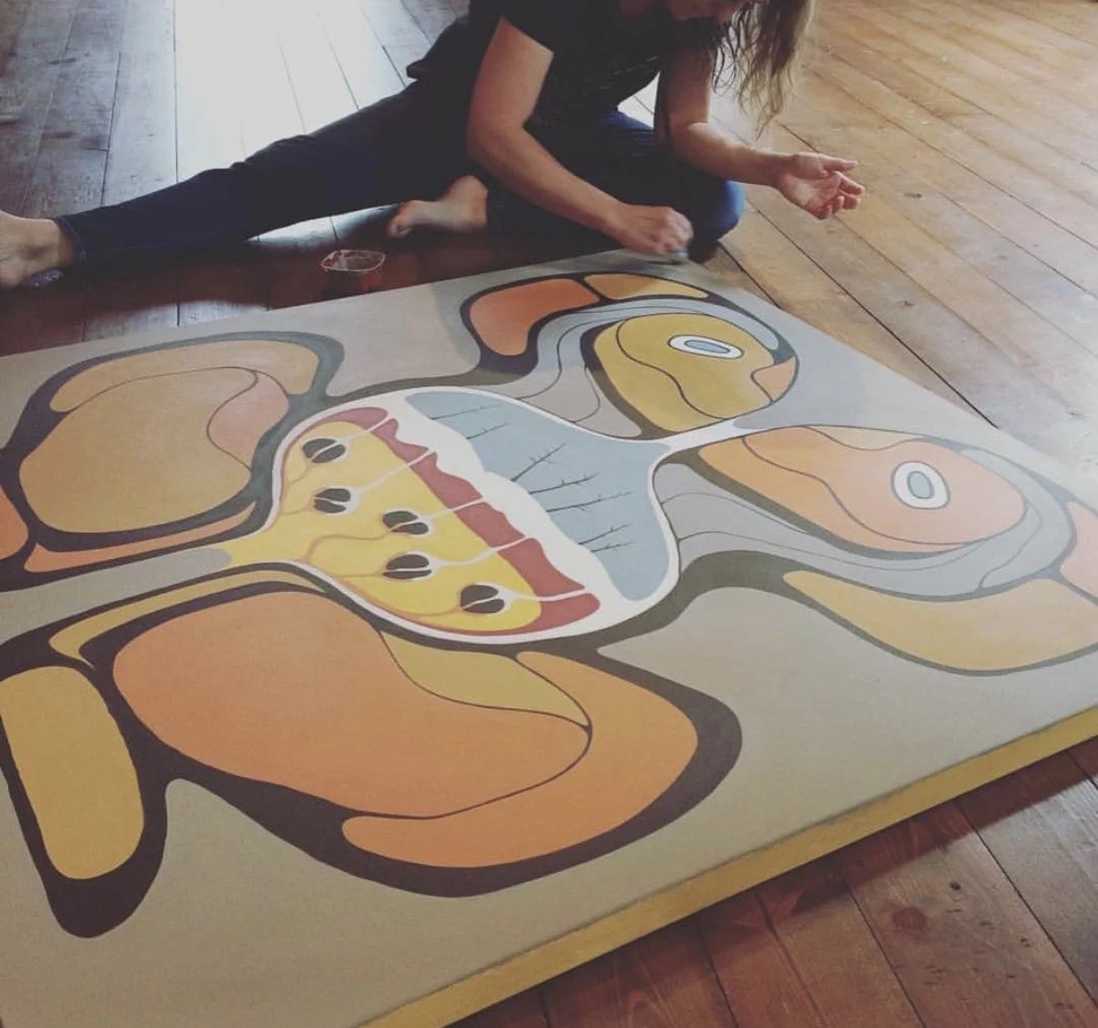 A person sitting on a wooden floor working on a large abstract painting with organic shapes and bold black outlines, featuring shades of orange, yellow, and gray.