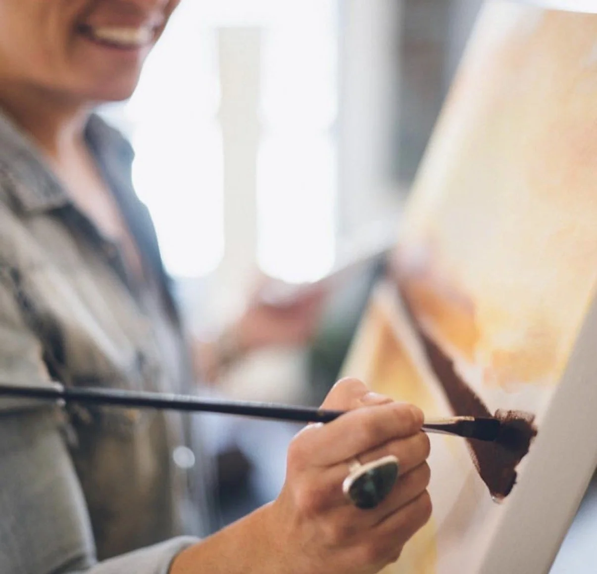 Person smiling while painting a landscape on a canvas with a paintbrush.