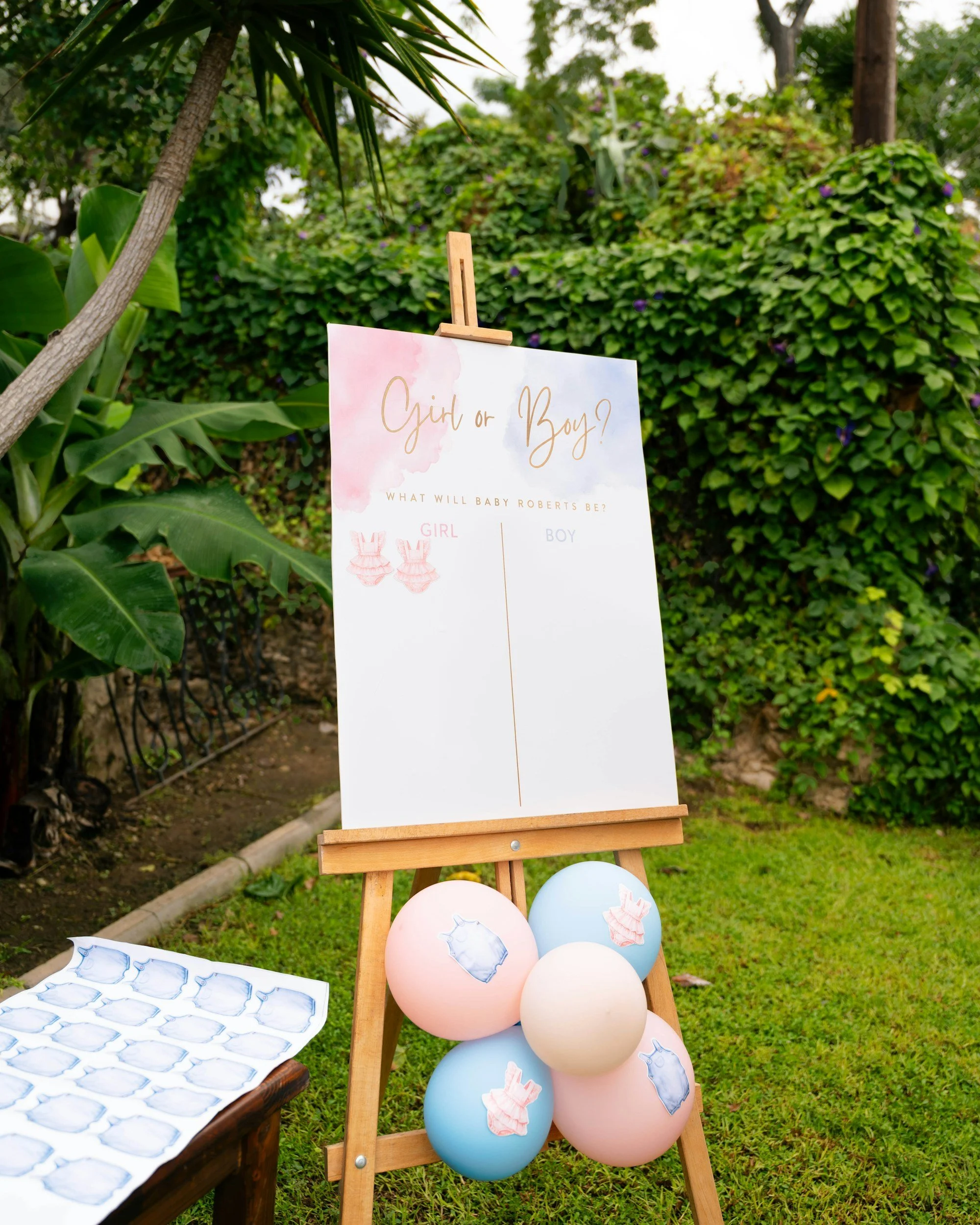 A decorated outdoor display for a gender reveal party with balloons in pink and blue, a whiteboard with 'Girl or Boy?' written on it, and scattered cutouts of baby clothes and bottles, set against green bushes and trees.