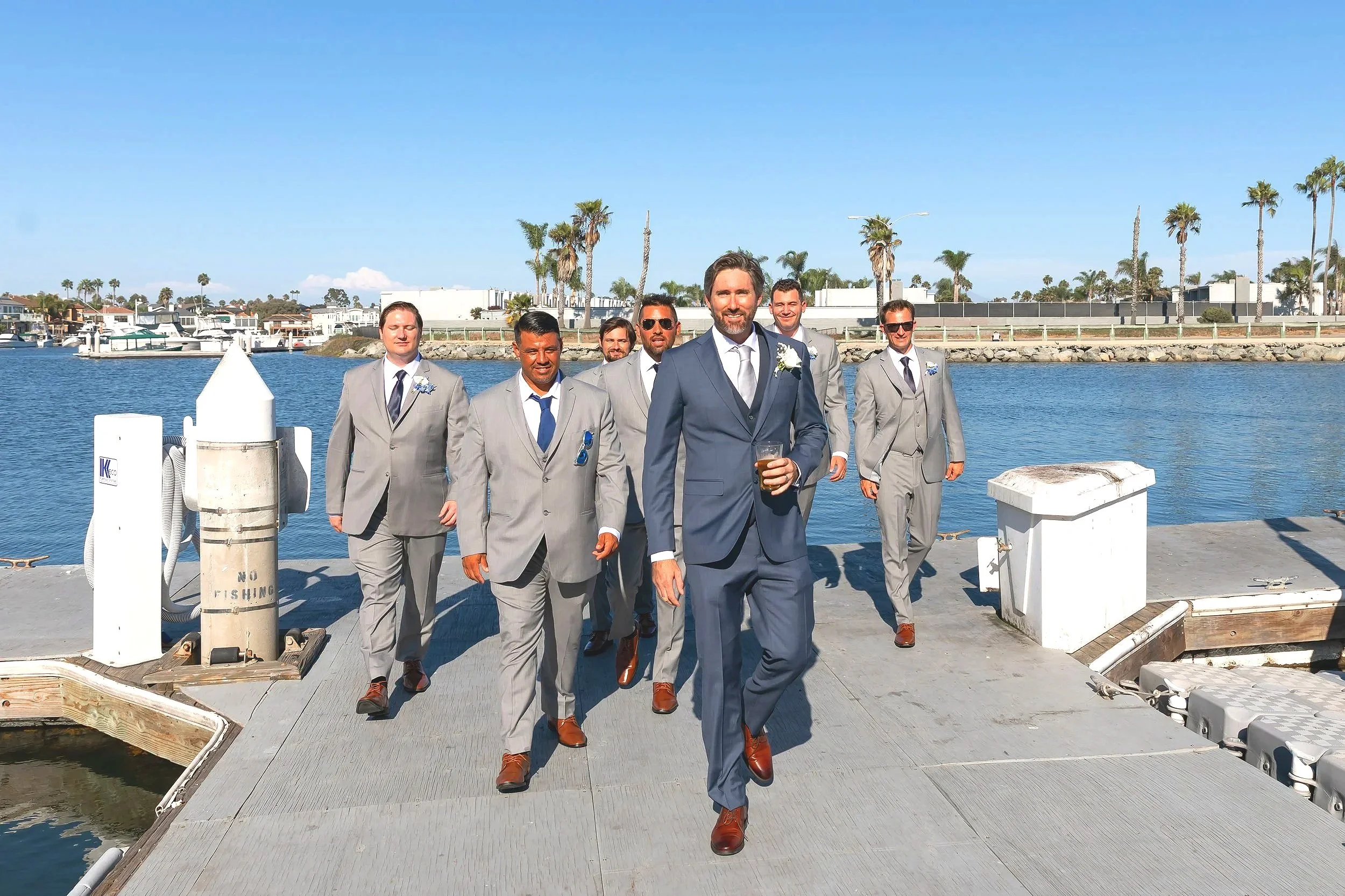 A group of seven men in gray suits and one man in a blue suit walk on a dock near the water, with boats and palm trees in the background.