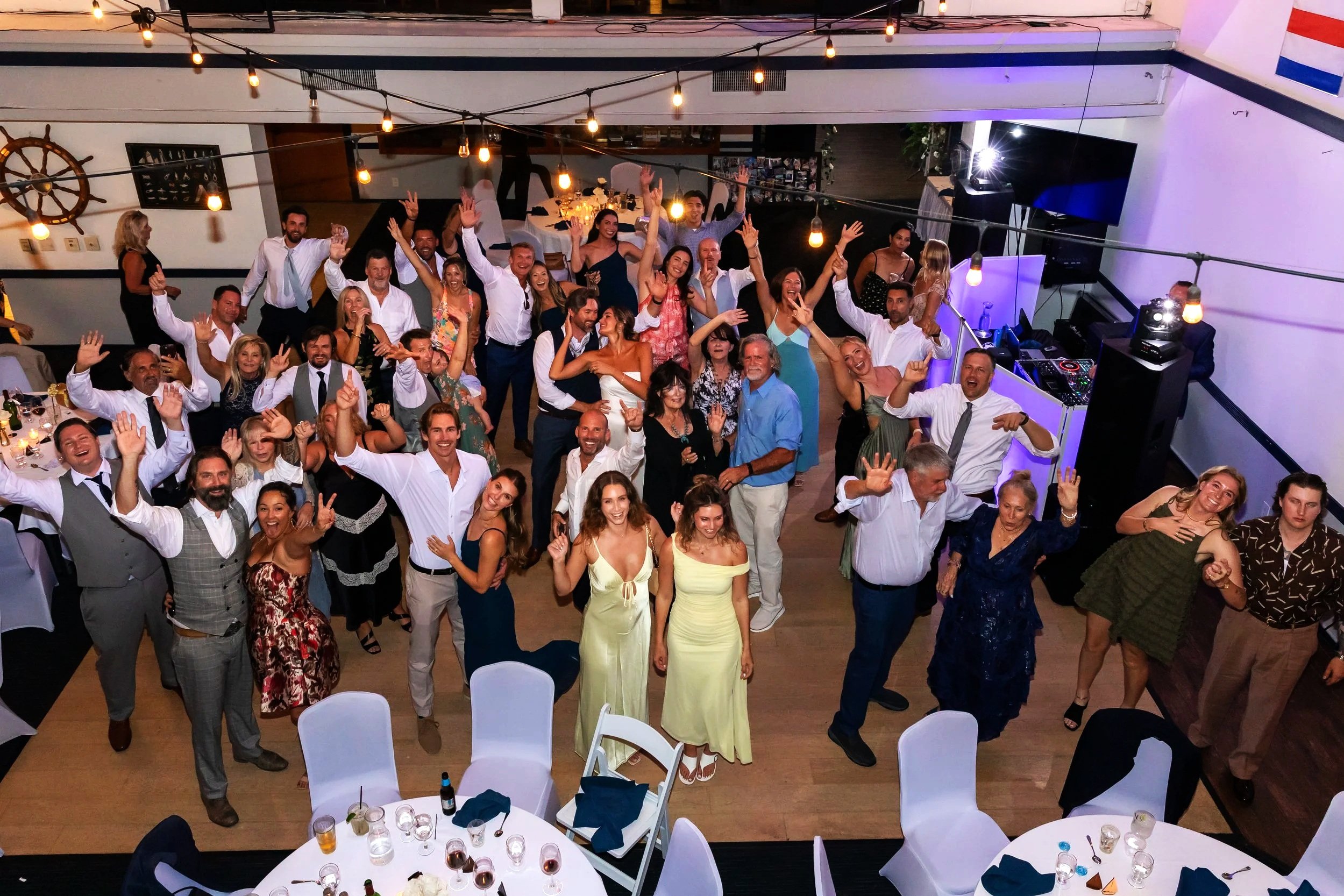 Group of people celebrating at a party, dancing and smiling, with tables set with glasses and drinks in a decorated indoor venue with hanging lights.