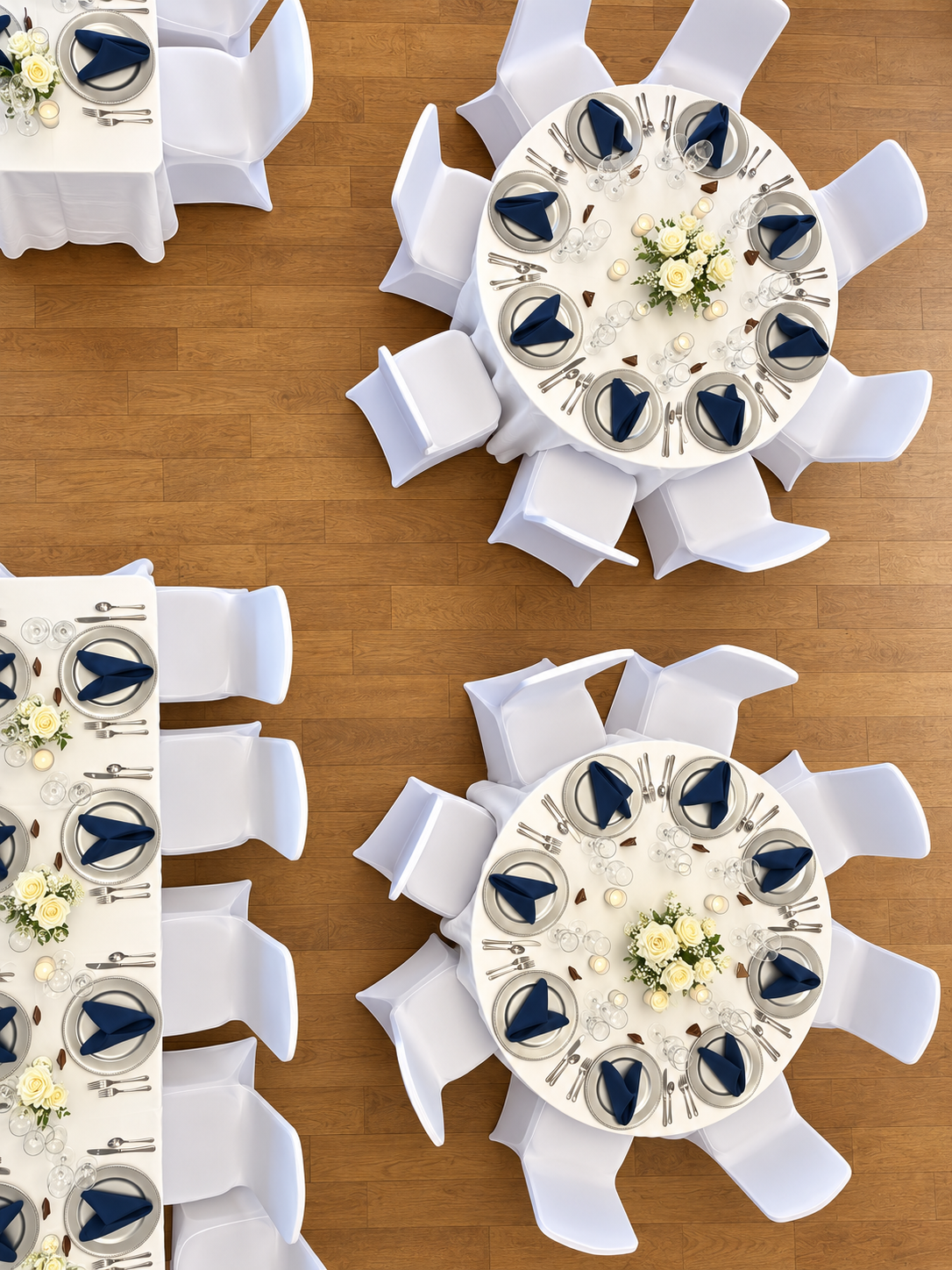 Overhead view of multiple round banquet tables with white tablecloths, floral centerpieces, and place settings including plates, silverware, wine glasses, and navy blue napkins, arranged on a wooden floor.