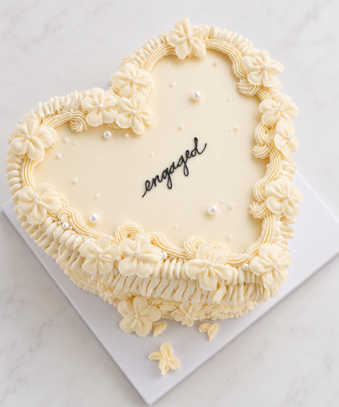 A heart-shaped wedding cake decorated with white flowers, pearls, and piped icing, with the word 'engaged' written in black on top.