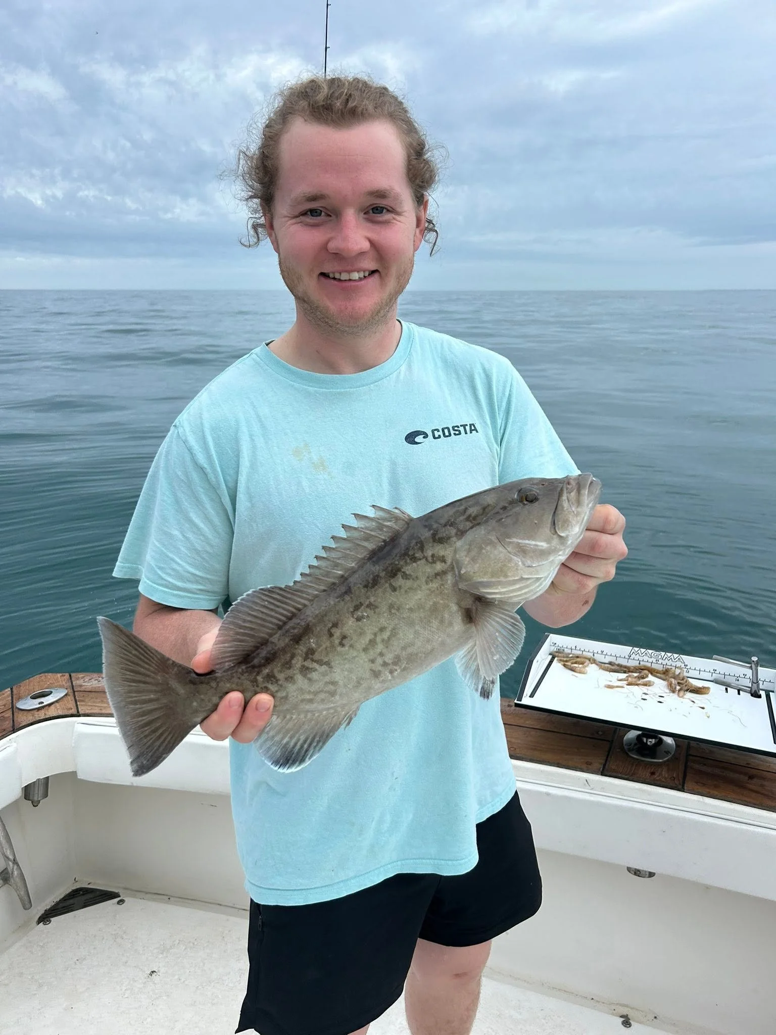 Smiling angler in a light blue shirt holding a large freshly caught fish aboard a fishing boat.