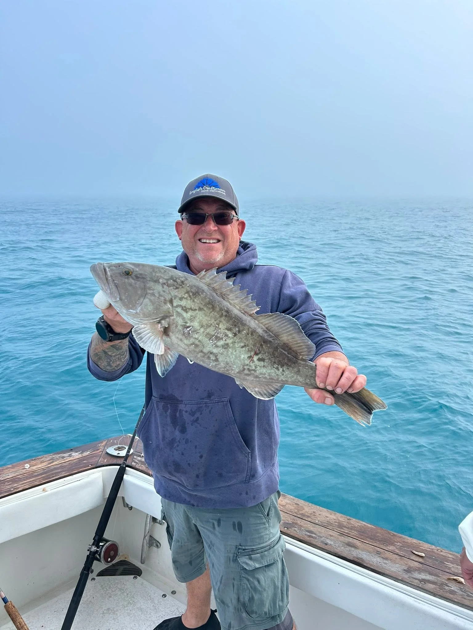 Smiling angler wearing a blue hoodie and sunglasses holding a large freshly caught fish aboard a fishing boat.