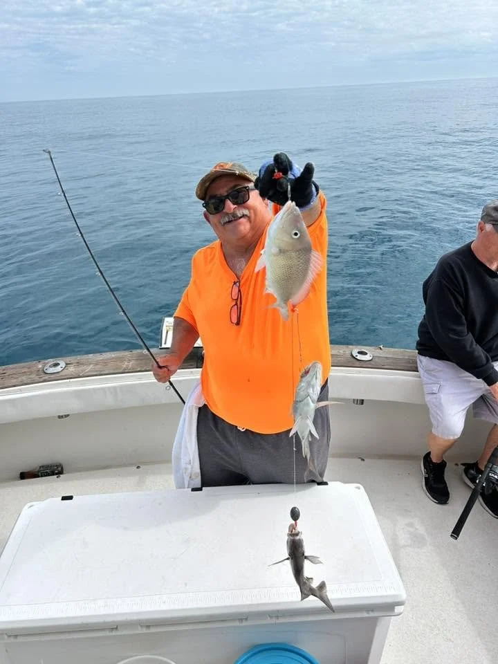 Smiling angler in a bright orange shirt holding up two freshly caught fish aboard a fishing boat.