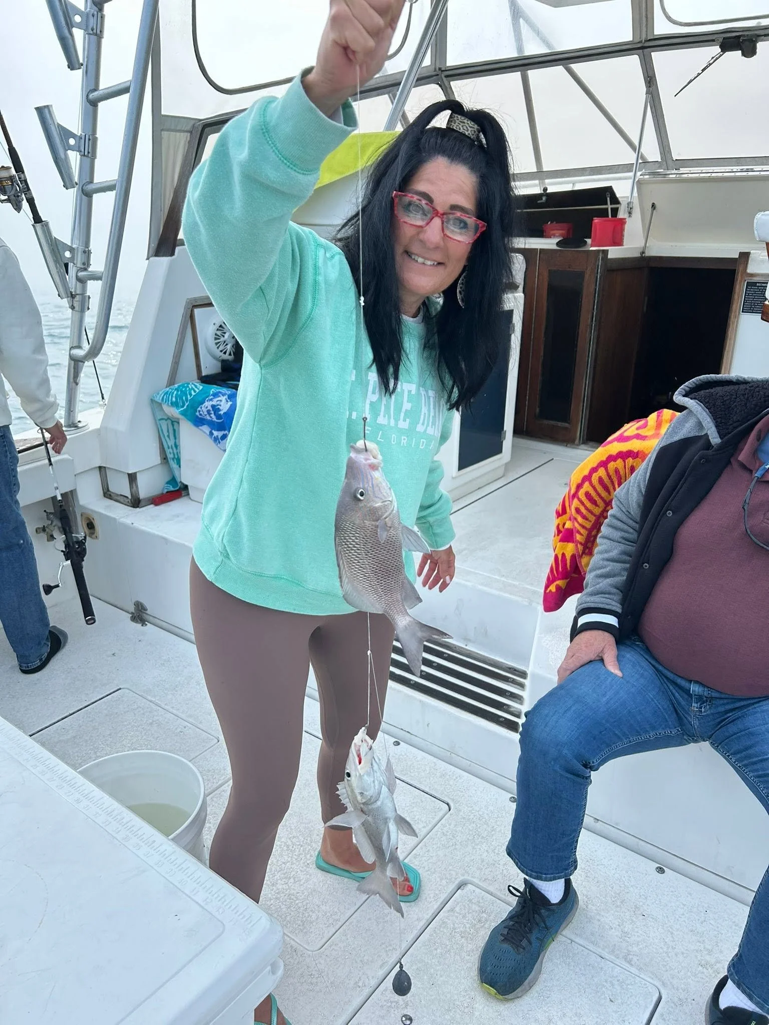 Smiling guest in a turquoise sweatshirt displaying two freshly caught fish aboard a fishing boat.