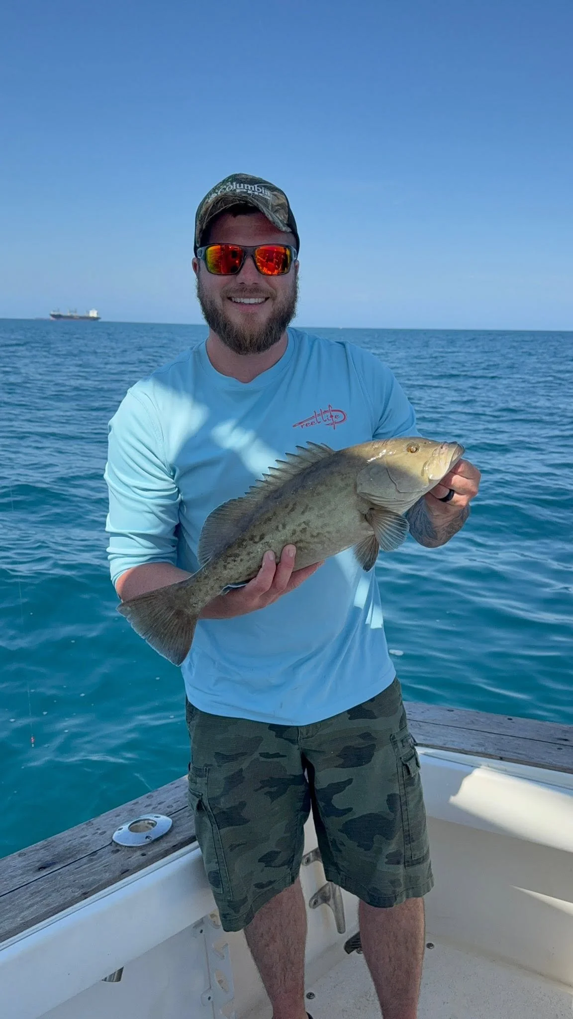 Angler wearing a light blue fishing shirt holding a large freshly caught fish aboard a boat on the Gulf of Mexico.