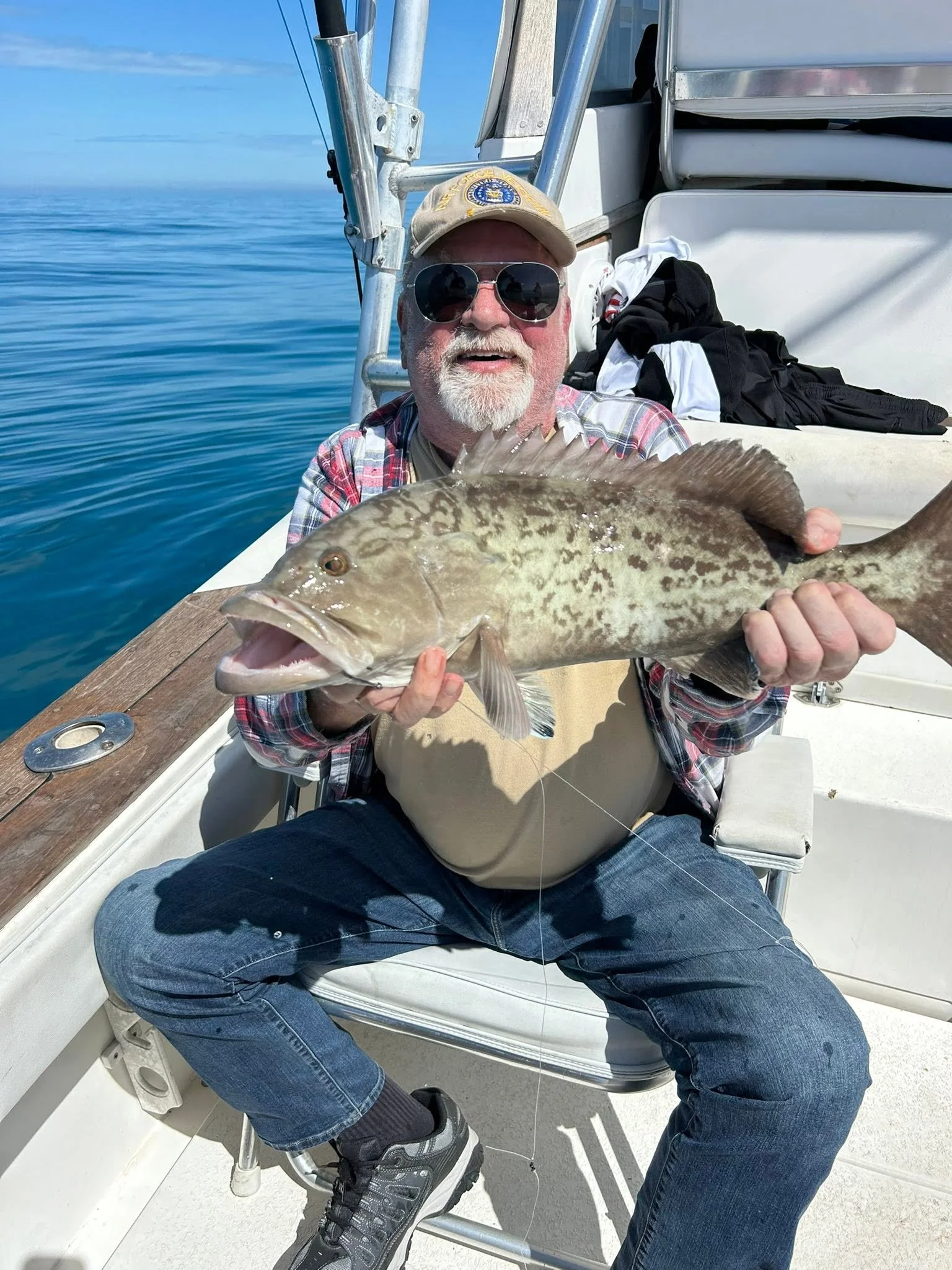 Bearded angler wearing sunglasses proudly holding a large spotted grouper aboard a fishing boat.