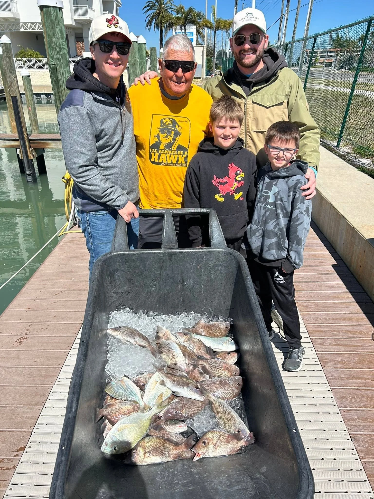 Two young anglers posing with family members behind a cart filled with freshly caught fish at the marina.