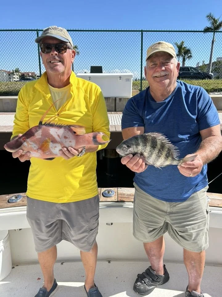 Two anglers holding freshly caught fish, one pink and orange and the other dark striped, aboard a fishing boat.