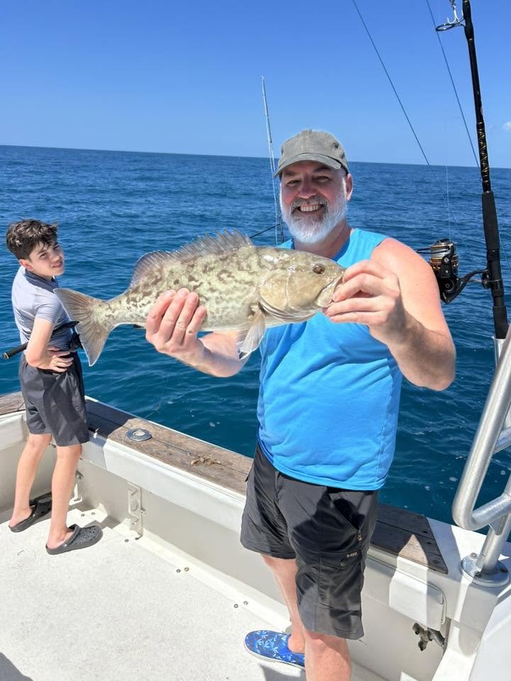 Smiling angler in a bright blue shirt holding a large spotted fish aboard a fishing boat, with a young guest nearby.